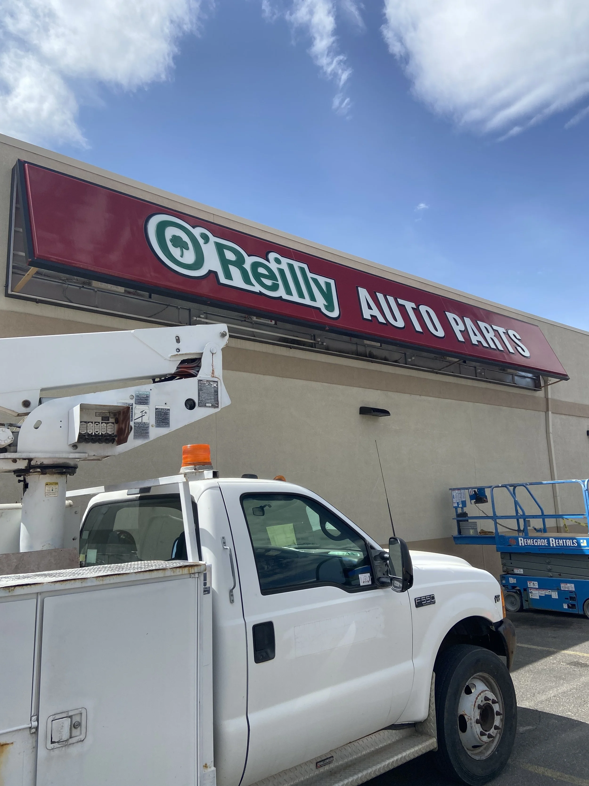 A white utility truck with a bucket lift is parked in front of a red sign for 'O'Reilly Auto Parts' store on a partly cloudy day.