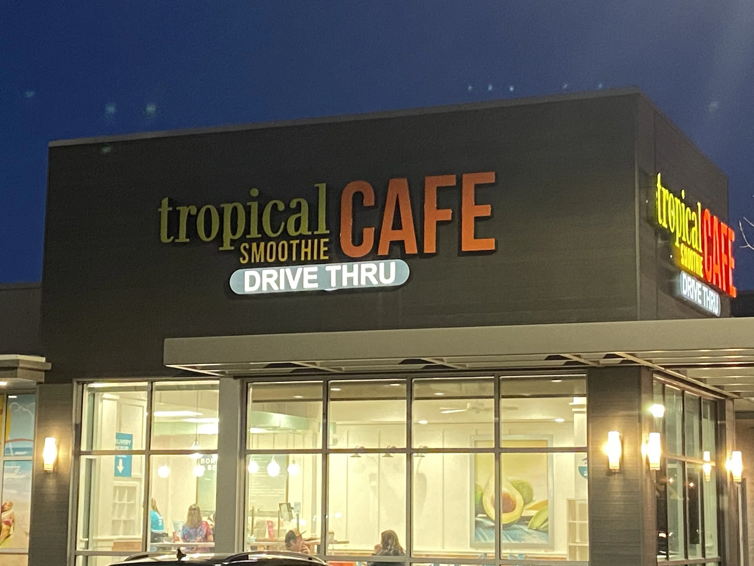 Nighttime exterior view of a Tropical Café with illuminated sign, featuring drive-thru, large glass windows showing people inside, and yellow lighting.