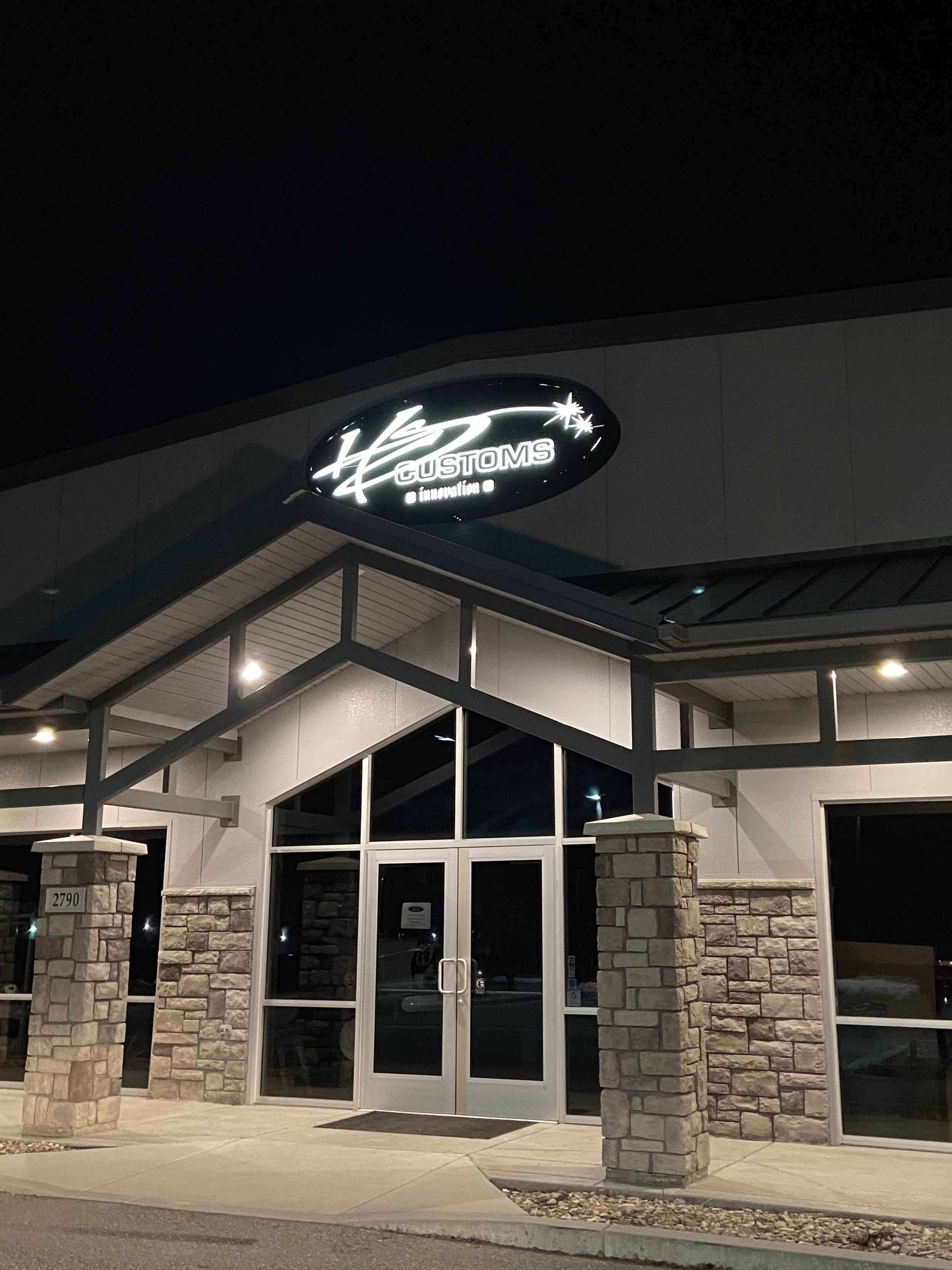 Night view of the entrance to a commercial building with a sign that reads 'LED CUSTOMS innovation.' The entrance features a glass door, brick columns, and a modern metal awning. Exterior lighting highlights the building's facade.