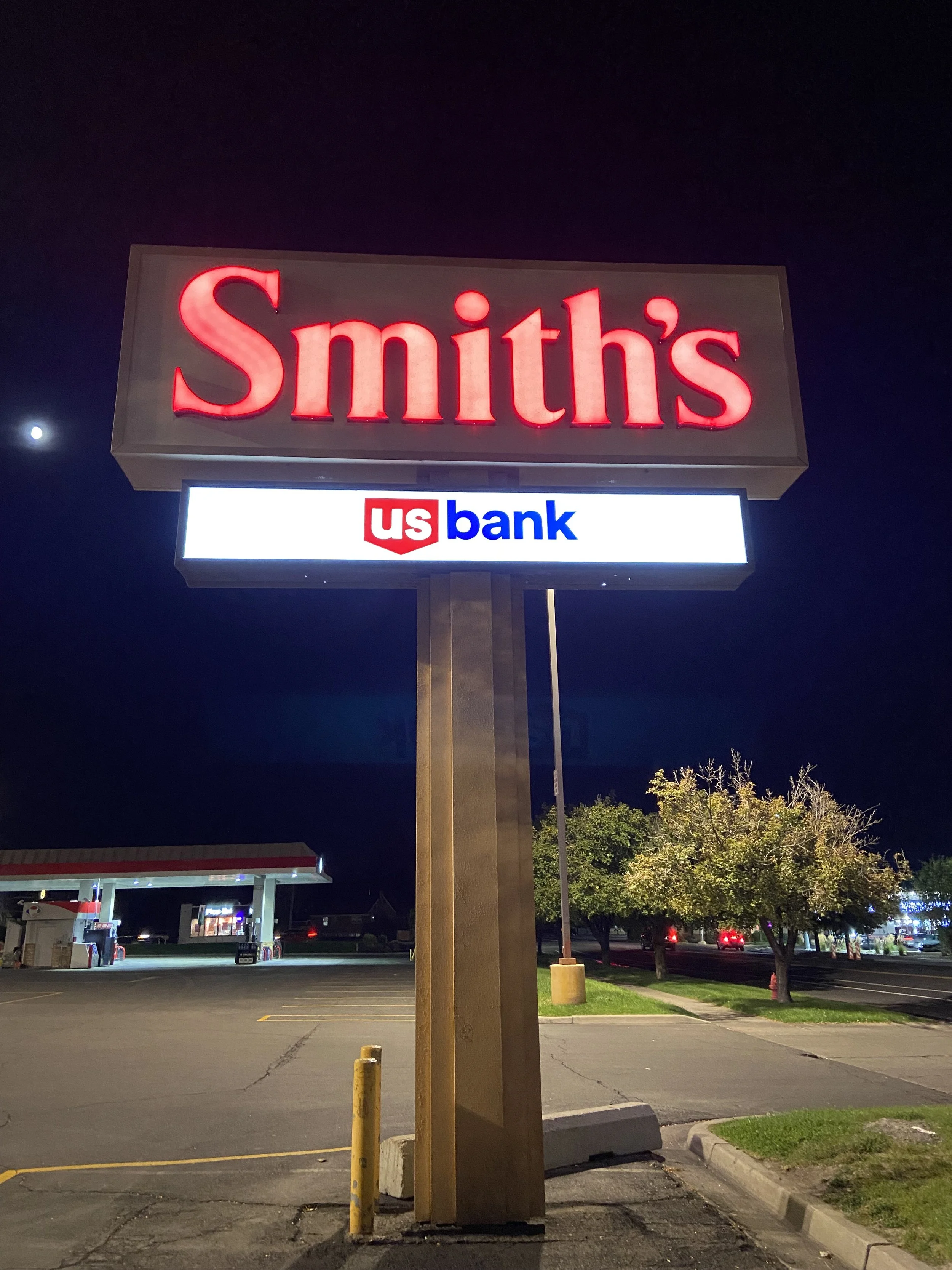 Nighttime scene of a parking lot with a large illuminated sign reading 'Smith's' in red, and a smaller sign below displaying 'US Bank' in red, white, and blue. The lot is mostly empty with a gas station visible in the background, some trees, and a clear sky with the moon visible.