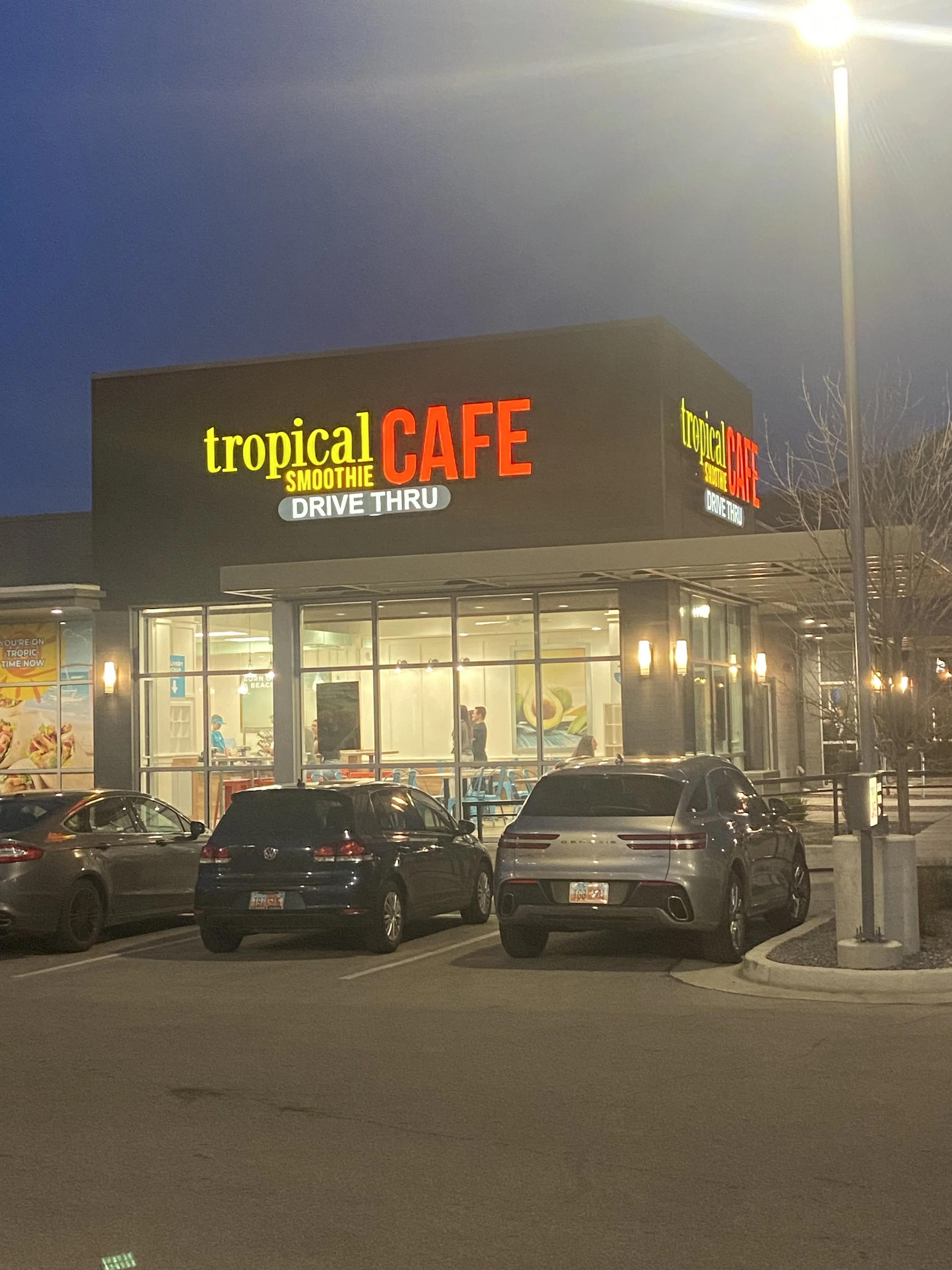 A parking lot in front of a Tropical Smoothie Cafe restaurant at dusk, with several parked cars and a lit sign on the building.