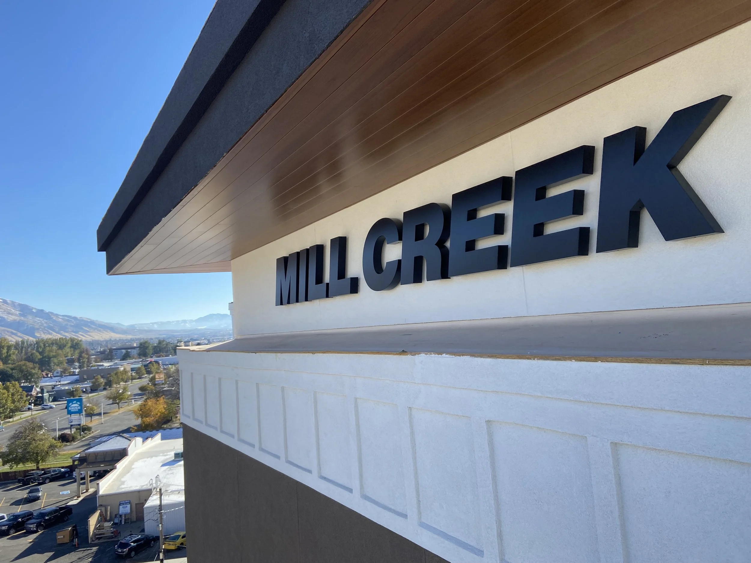 Exterior view of a building with a sign that reads 'Milk Créek' on the side. The building has white and dark gray walls with a wooden accent. In the background, there are trees, roads, and distant mountains under a clear blue sky.
