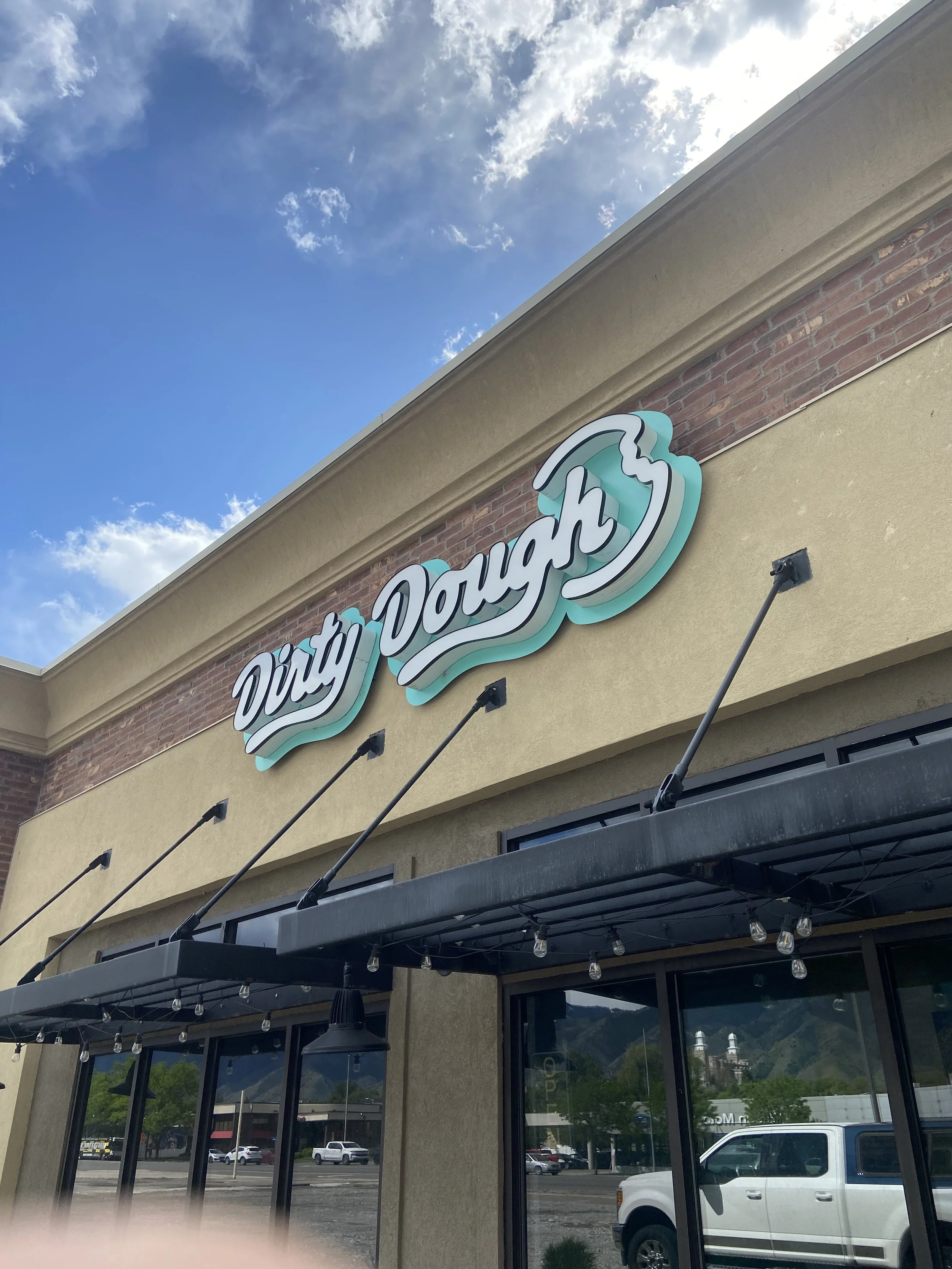 Storefront with a sign that says 'Dirty Dough' above the entrance, with large windows and outdoor lighting fixtures, cars parked in front, and partly cloudy sky overhead.