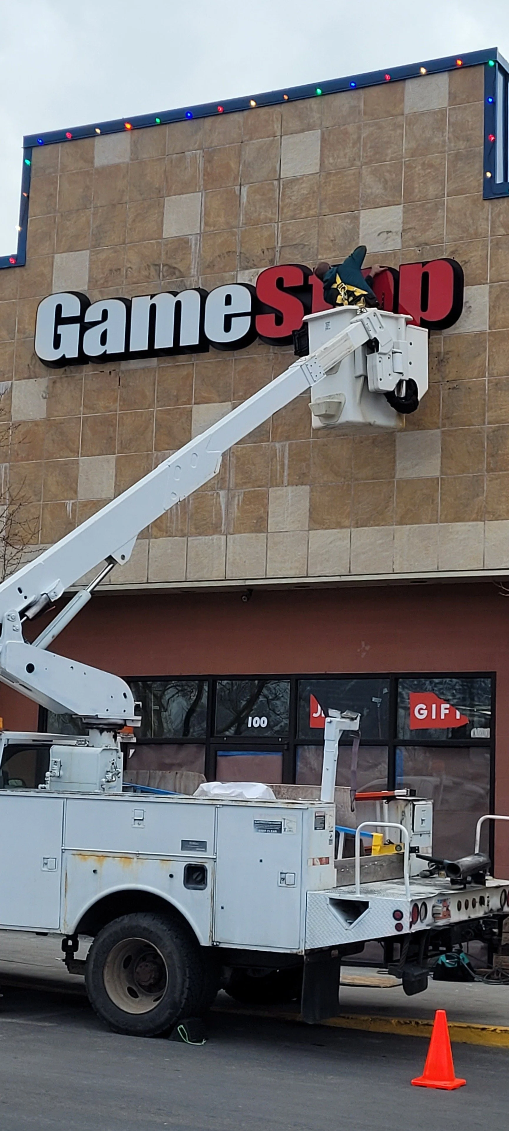 A worker on a cherry picker installing or repairing the sign of 'GameStop' store on the building's exterior.