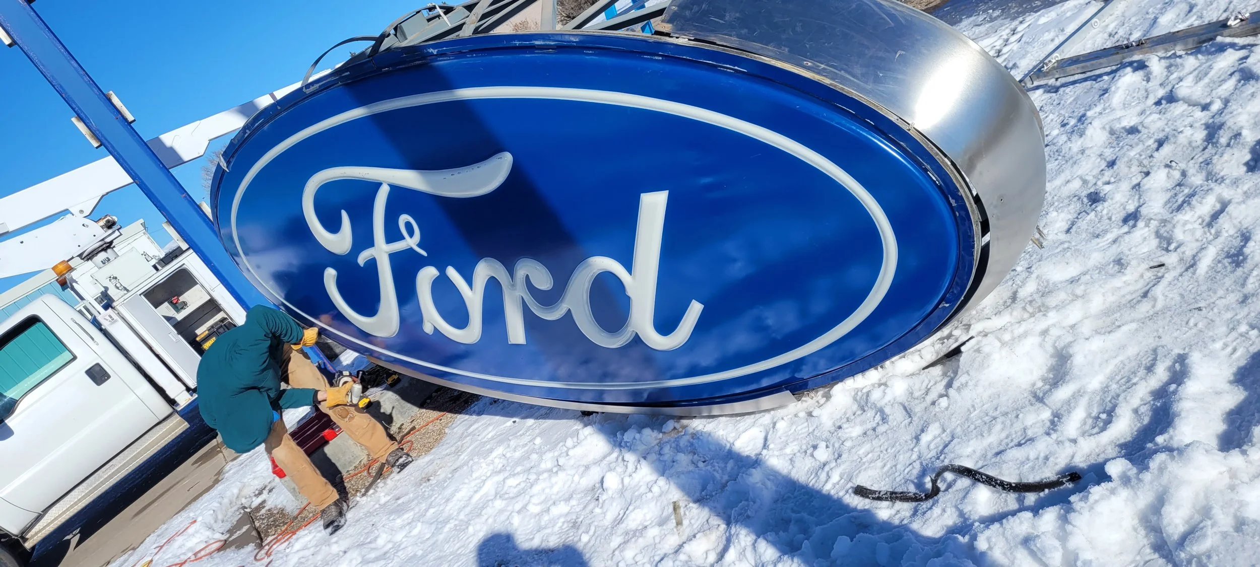 A large outdoor sign with the Ford logo being installed in a snowy area, with workers working on the sign and a truck nearby.