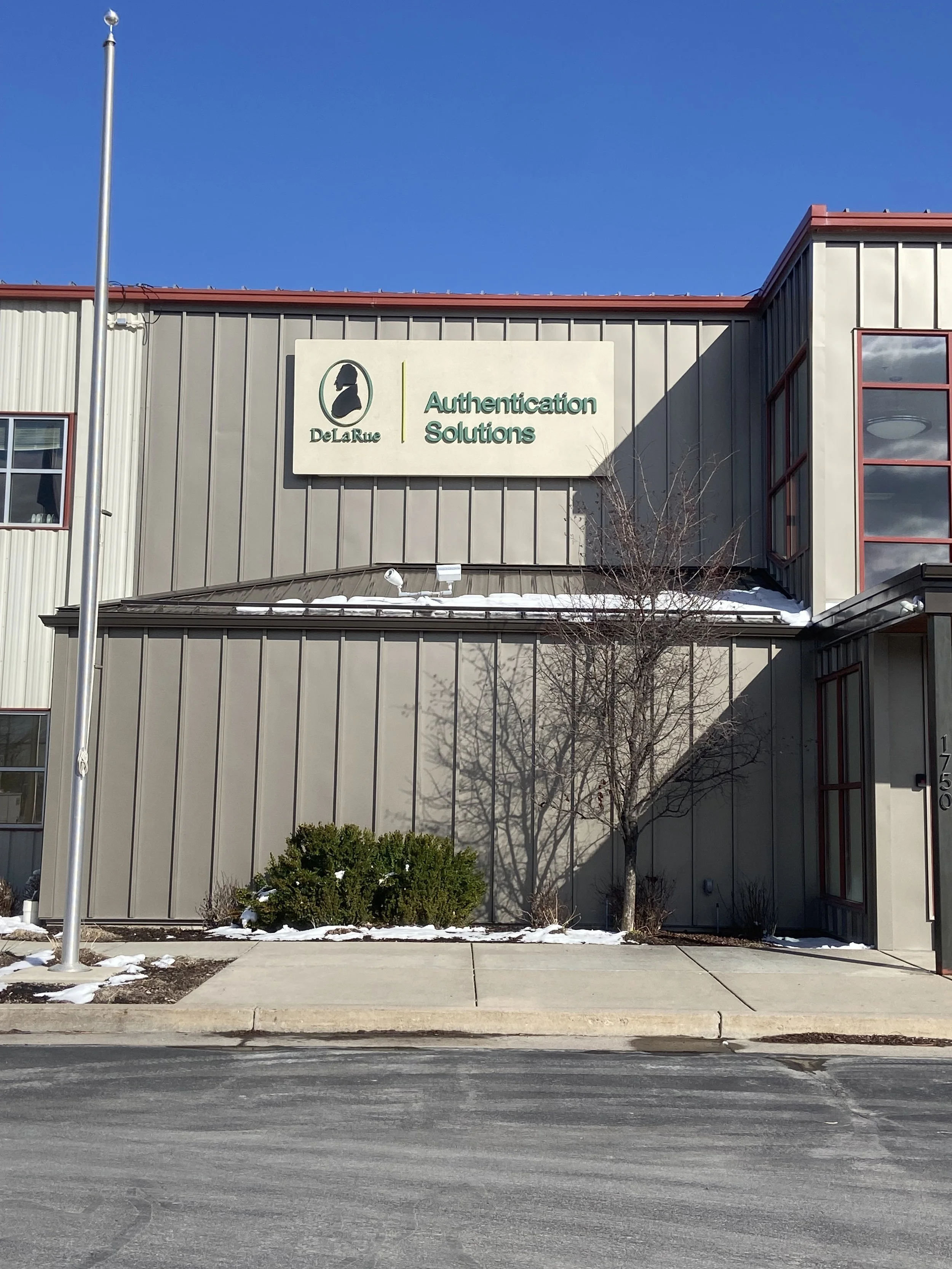 Front of a building with a sign that reads 'De La Rue Authentication Solutions,' a flagpole with no flag, a leafless tree, some bushes, snow on the ground, and a clear blue sky.