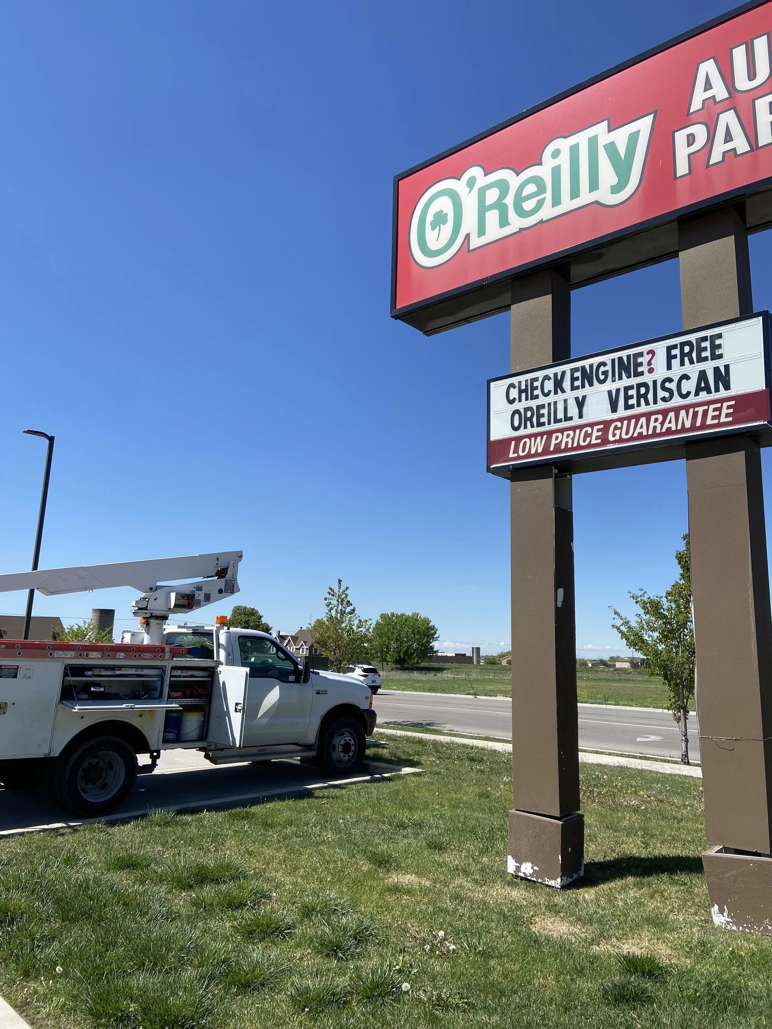A large shopping plaza sign for O'Reilly Auto Parts with additional message boards advertising check engine service, free O'Reilly Veriscans, and a low-price guarantee. There is a utility vehicle with an aerial lift parked underneath the sign, and a street with a few trees and small buildings in the background.