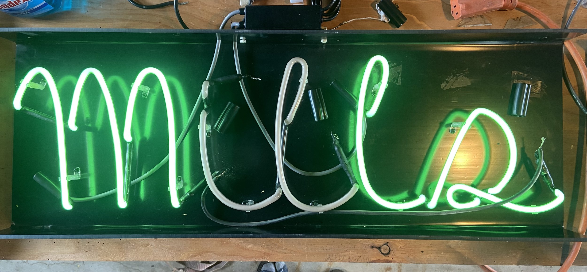 A neon sign with the word 'Mills' written in green cursive letters, mounted on a black rectangular background board.
