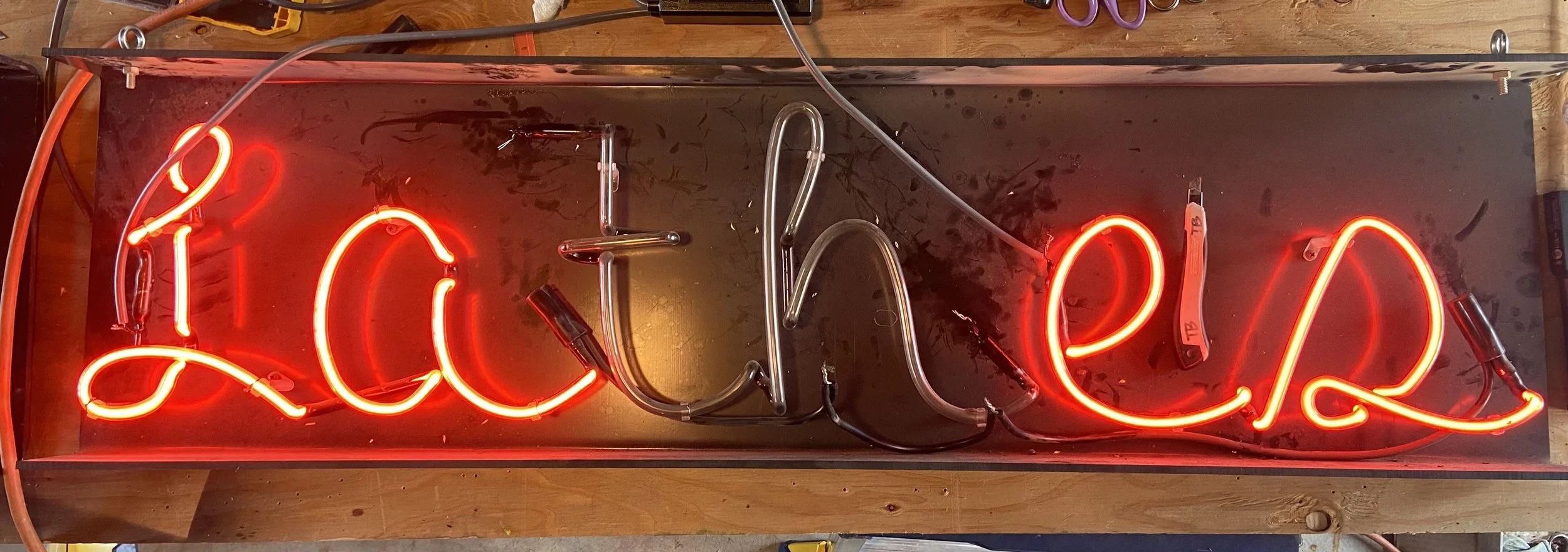 A neon sign that spells out "Lathes" in red cursive letters, mounted on a black rectangular background, with electrical wires and components visible behind it.