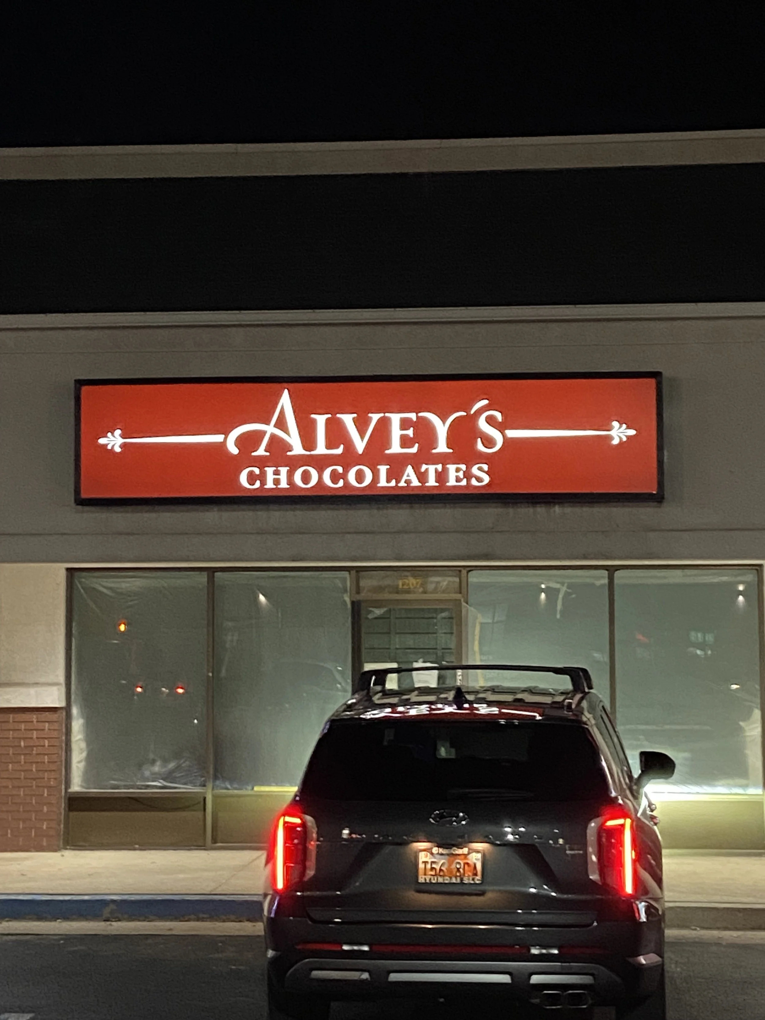 A black SUV parked in front of a store called Alvey's Chocolates at night, with a red illuminated sign.