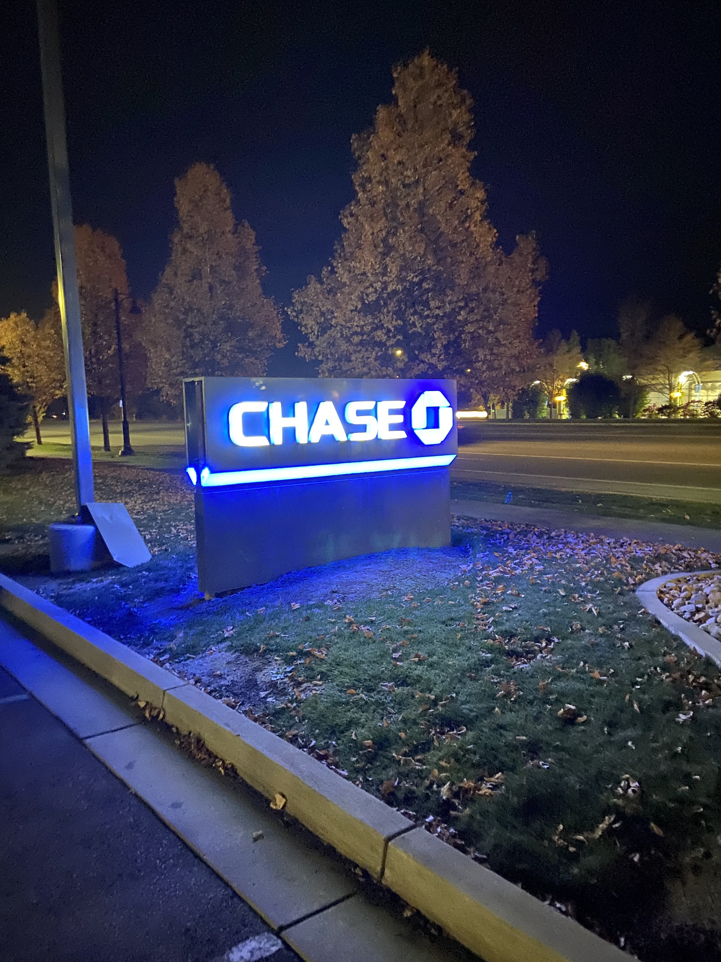 Nighttime view of a Chase bank sign with illuminated blue and white logo, trees with autumn leaves, and a street with fallen leaves on the ground.