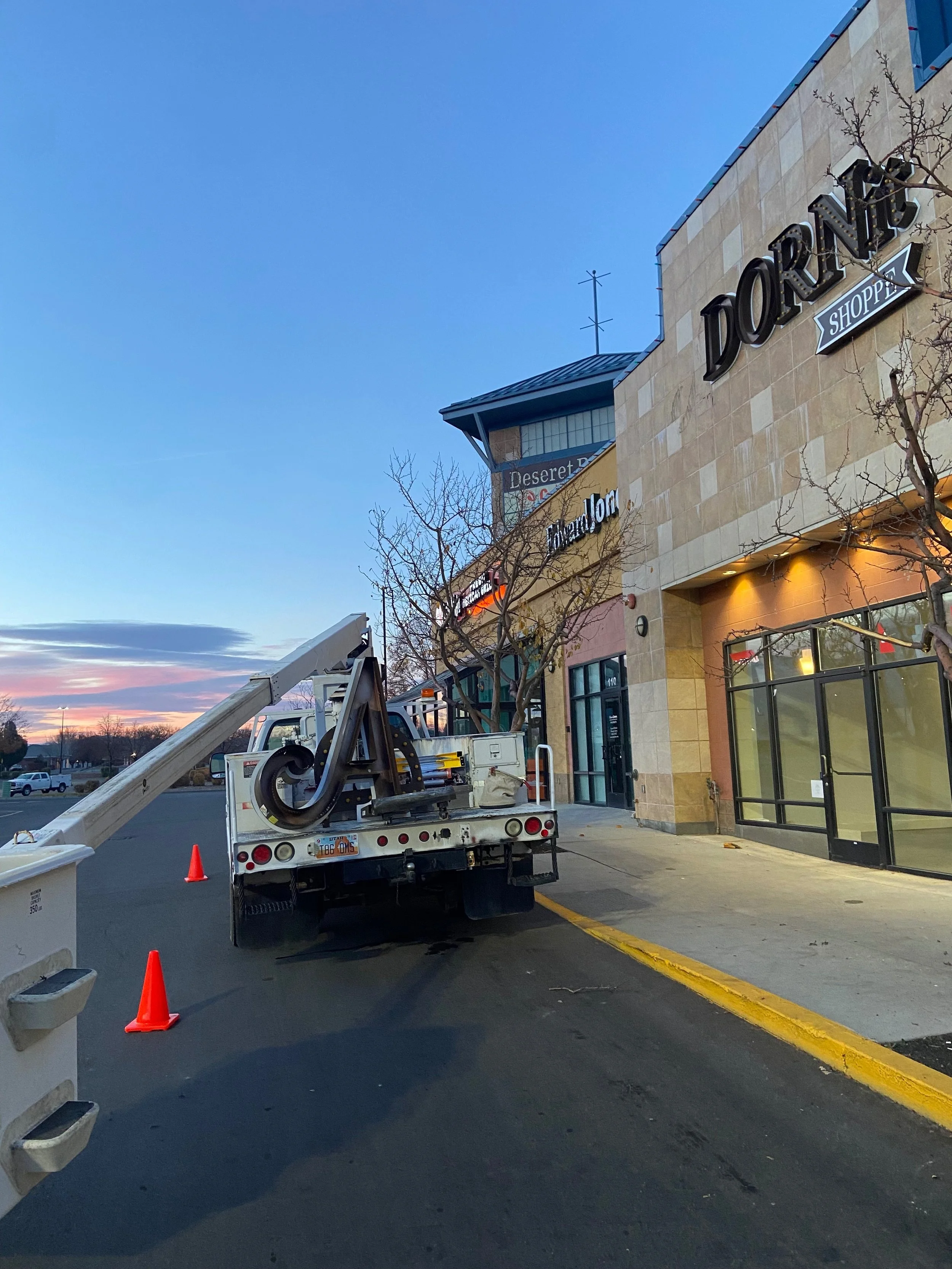 A parking lot outside a shopping center with a truck that has a lift or crane, orange traffic cones, and under the sky at sunset.