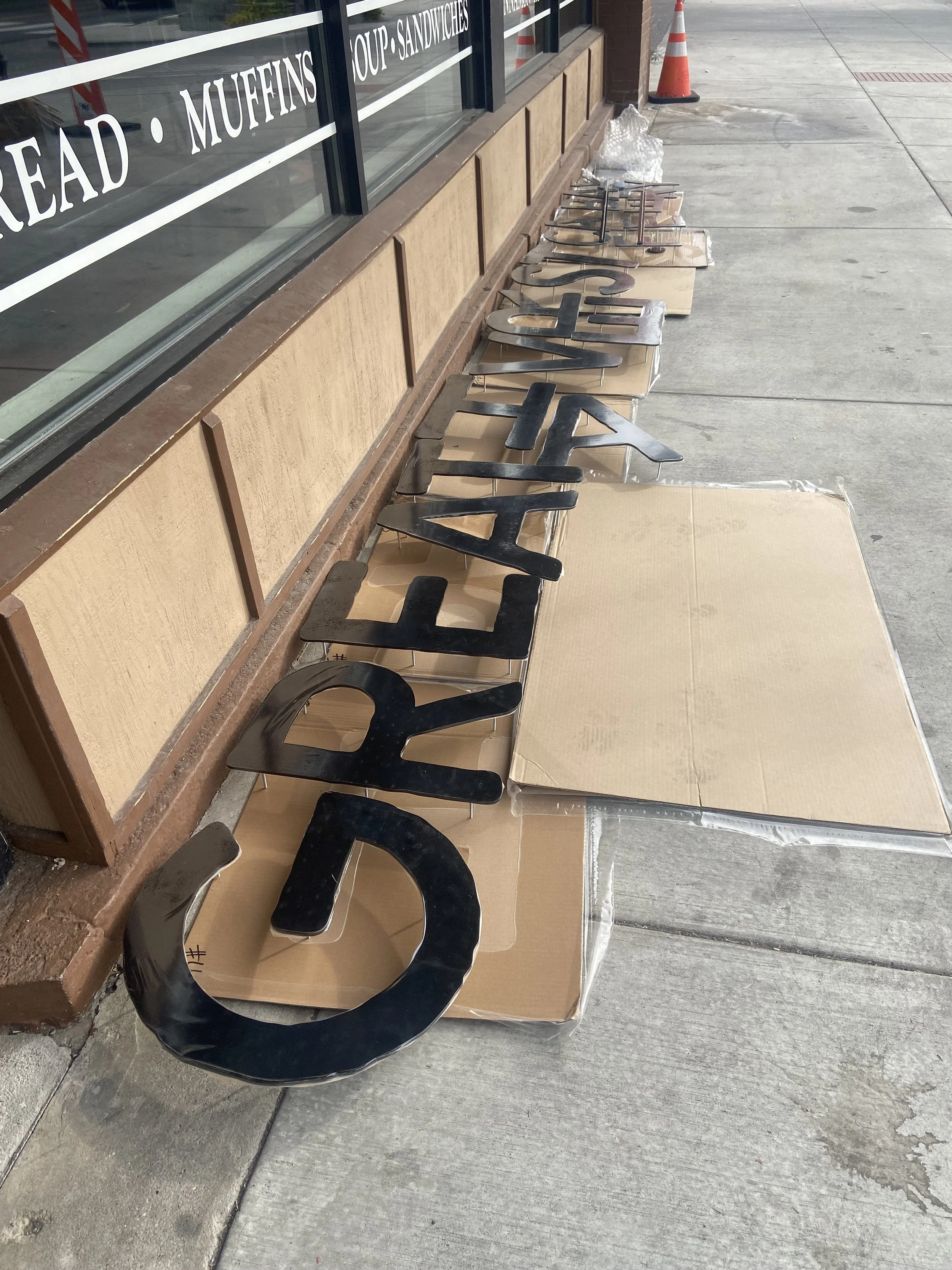 Black letters spelling out various words arranged on cardboard on the sidewalk outside a store.