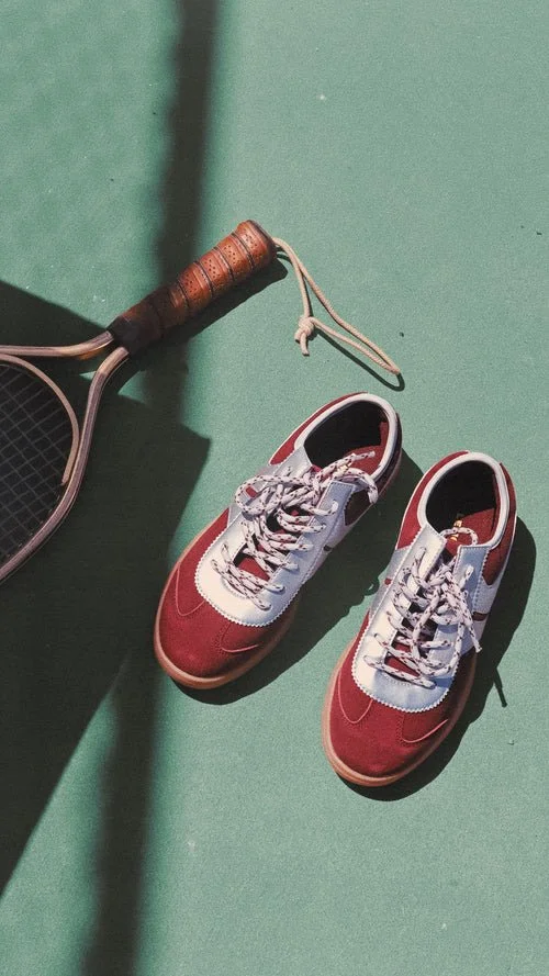 A pair of red and white sneakers and a tennis racket with a wooden handle on a green tennis court.