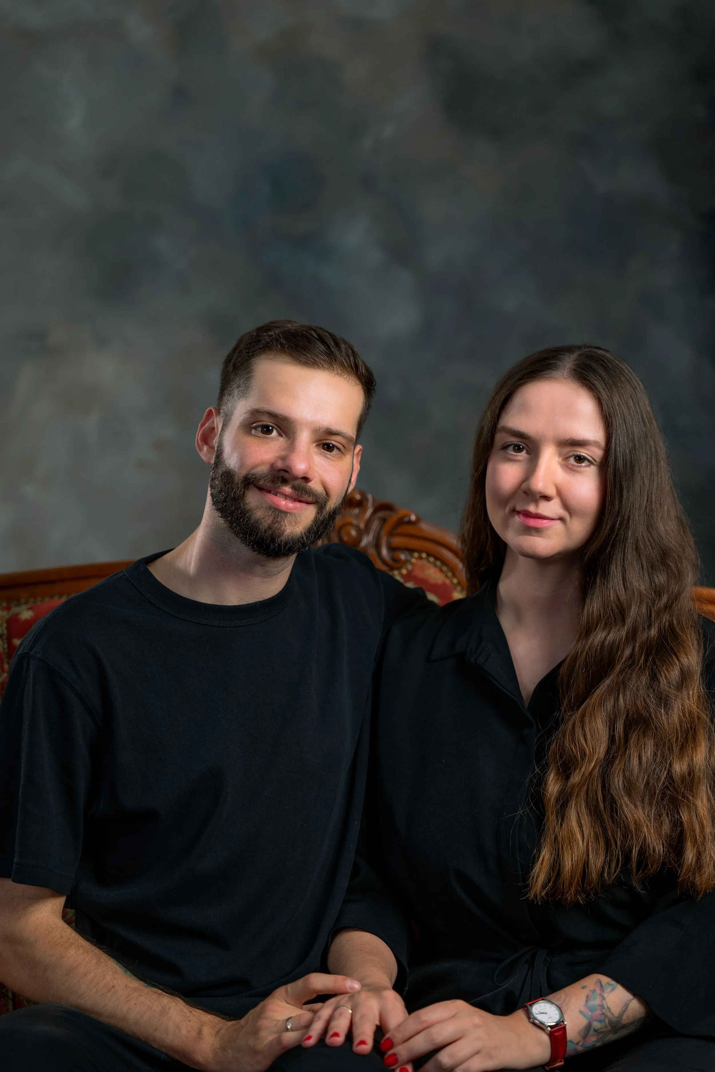 Ein Mann mit Bart und eine Frau mit langen braunen Haaren sitzen nebeneinander auf einem roten Polster mit geschnitztem Holzrahmen, vor einem dunklen, künstlerisch gestalteten Hintergrund. LoCrew starten ihr Podcast "Ungefragte Trauzeugen"