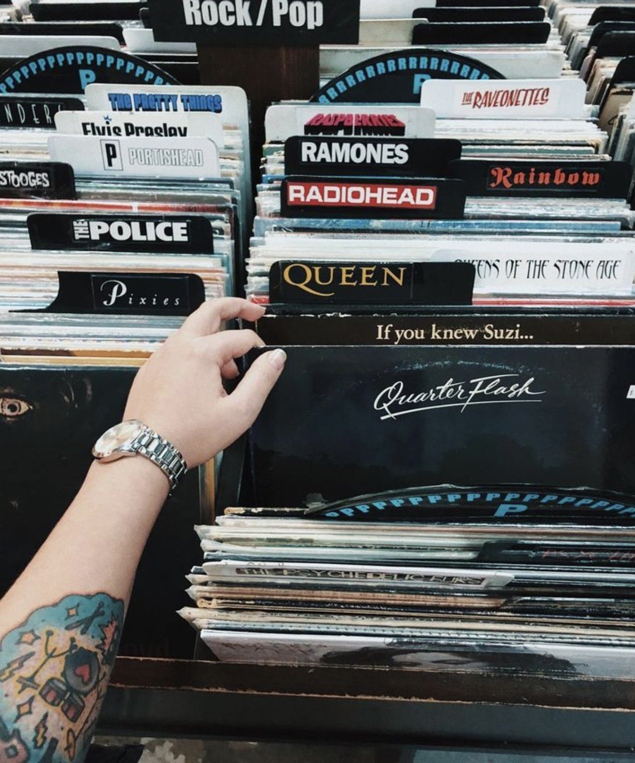 Person blättert durch Schallplatten in einer Rock/Pop-Abteilung eines Plattenladens. Verschiedene Bandnamen sind auf den Trennwänden sichtbar, einschließlich Queen, Radiohead, und Ramones. Eine tätowierte Hand greift nach einer Platte von Quarterflas