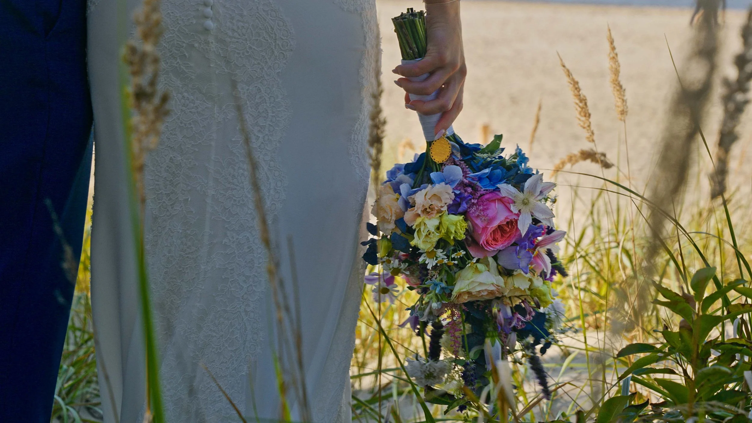 Ein Brautpaar bei der Hochzeit, die Braut hält einen bunten Blumenstrauß in der Hand, im Hintergrund ist Wasser und teilweise Natur zu sehen.