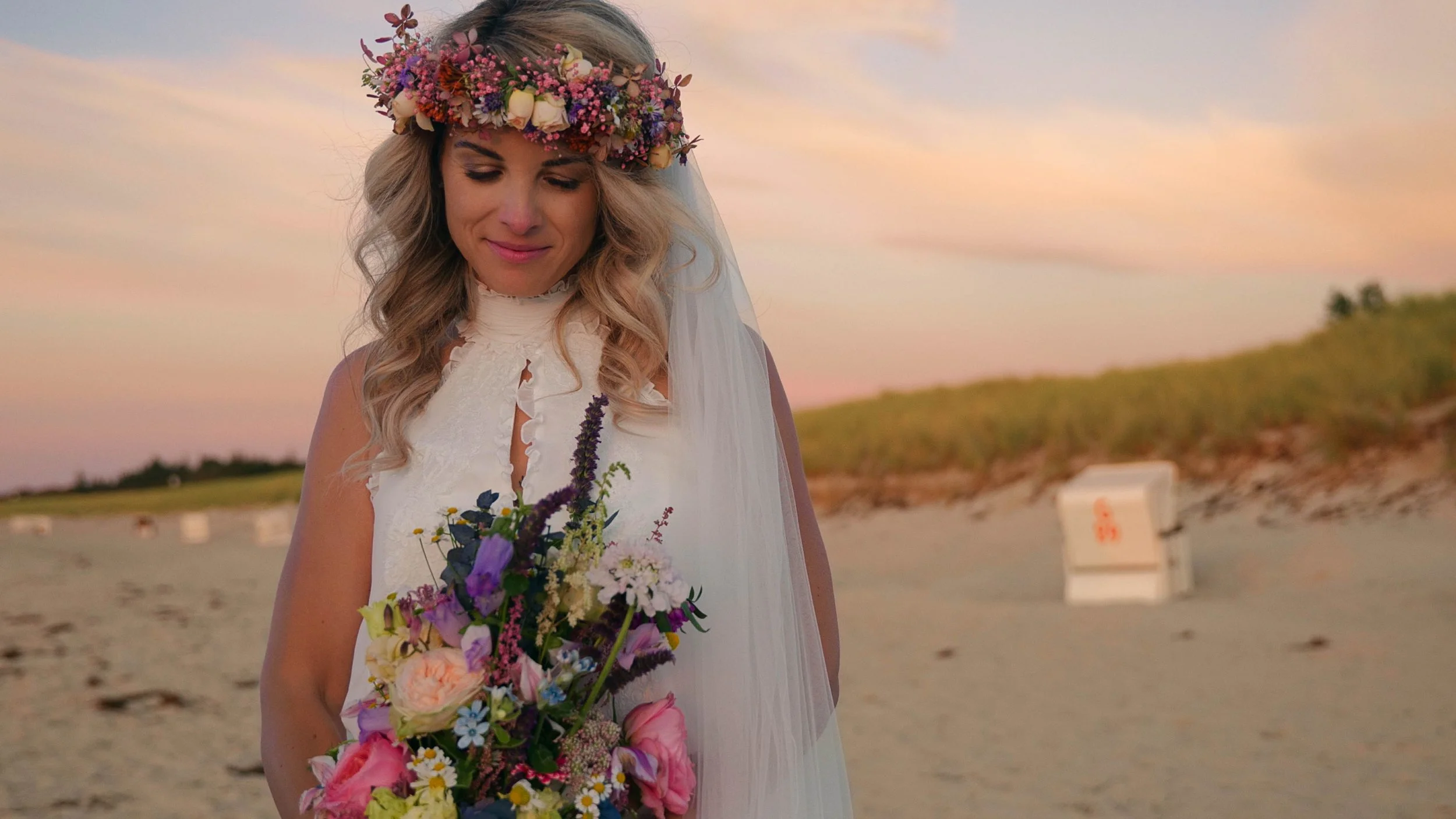 Eine Frau trägt ein weißes Brautkleid, einen Blumenkranz im Haar und einen Schleier auf einem Strand mit Sonnenuntergang im Hintergrund, hält einen bunten Blumenstrauß.