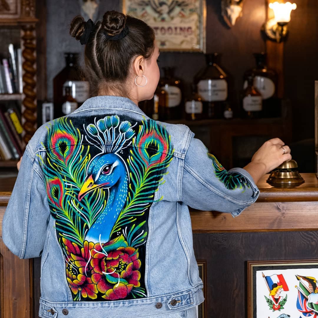 Frau mit Jeansjacke, Pfauendesign auf Rücken, in einem rustikalen Innenraum, Bücherregal und Glocke im Hintergrund.