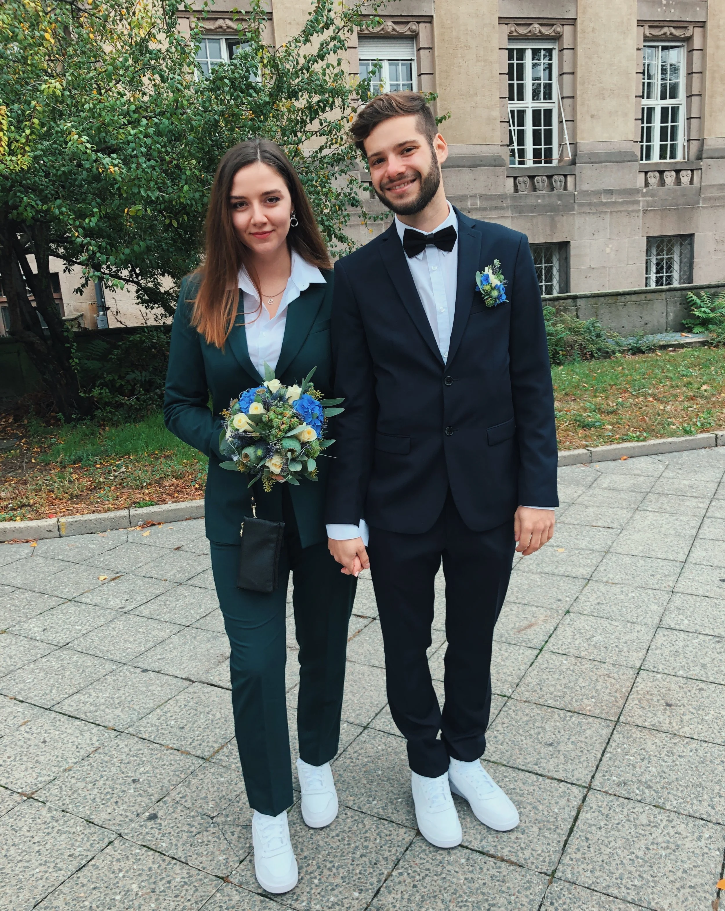 Brautpaar steht vor dem Standesamt. Die Frau trägt einen grünen Anzug und hält einen Blumenstrauß. Der Mann trägt einen dunkelblauen Anzug mit einer Fliege. Beide tragen weiße Schuhe und halten Händchen.