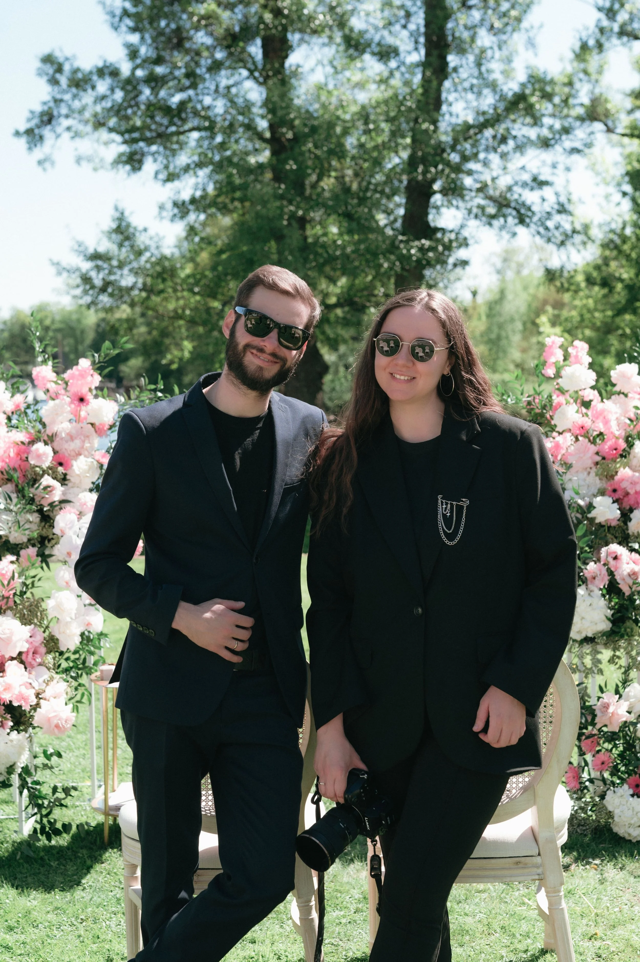Nico und Sofia - Hochzeitsvideografen LoCrew stehen im Freien vor dem Traubogen, beide tragen schwarze Anzüge und Sonnenbrillen, Sofia hält eine Kamera, es ist sonnig.