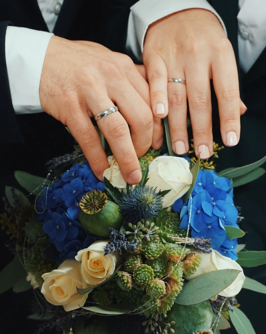 Zwei Hände mit Eheringen und einem Blumenstrauß; weiße Rosen und blaue Hortensien sind sichtbar.