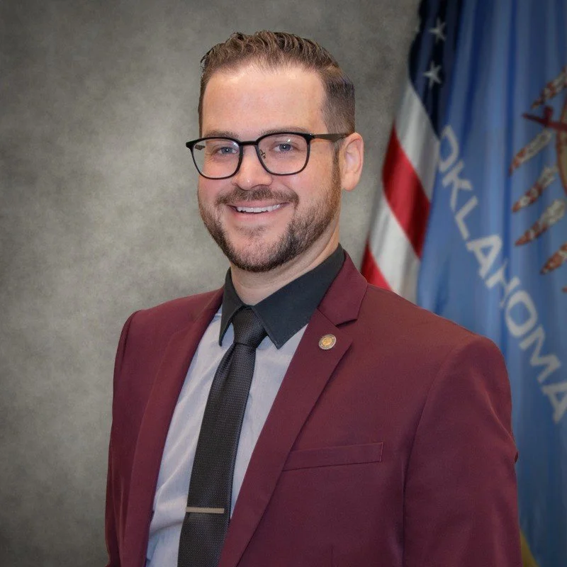 A man with glasses, wearing a maroon blazer, grey shirt, and black tie, smiling in front of an Oklahoma state flag.