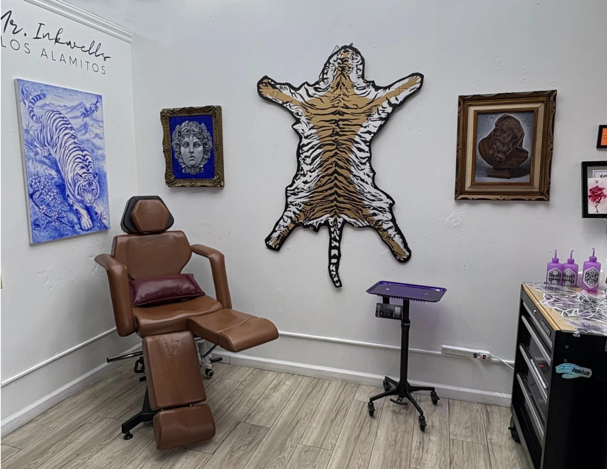 Art gallery wall with paintings of a tiger, a gemstone, and a reclining figure, and a cutout of a tiger, with chairs and a small table in a room.