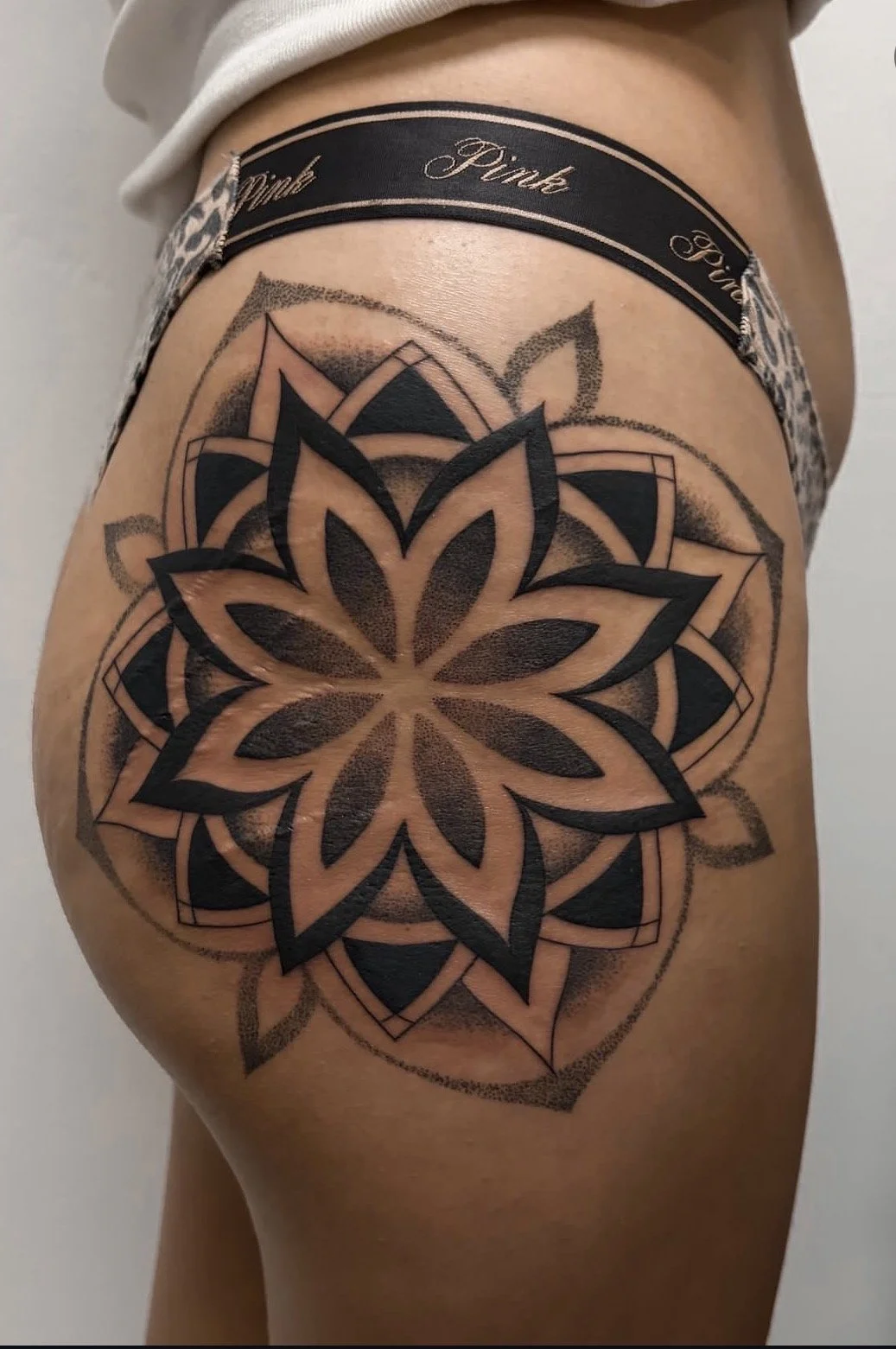 A person's upper thigh with a large, intricate black tattoo of a geometric floral mandala design, with layers of petals and concentric shapes. The person is wearing a black waistband with the words "Pink" and "Fun" written on it.