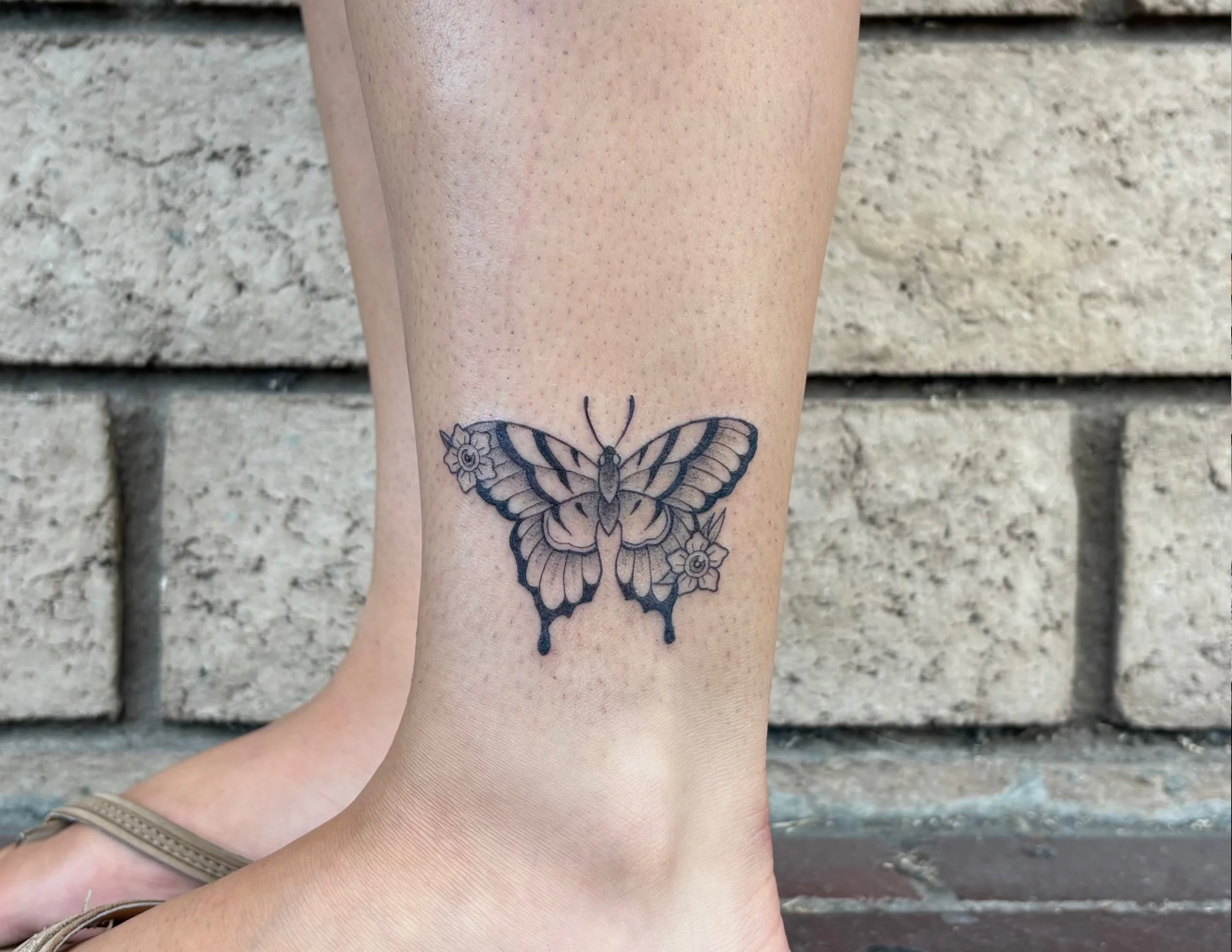 Close-up of a person's lower leg with a butterfly tattoo below the knee, standing against a brick wall.
