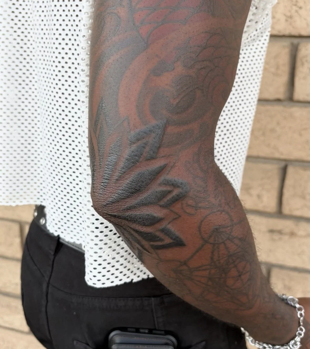 Close-up of a person's arm with a detailed black and red tattoo design, wearing a white perforated shirt and black pants, with a brick wall background.