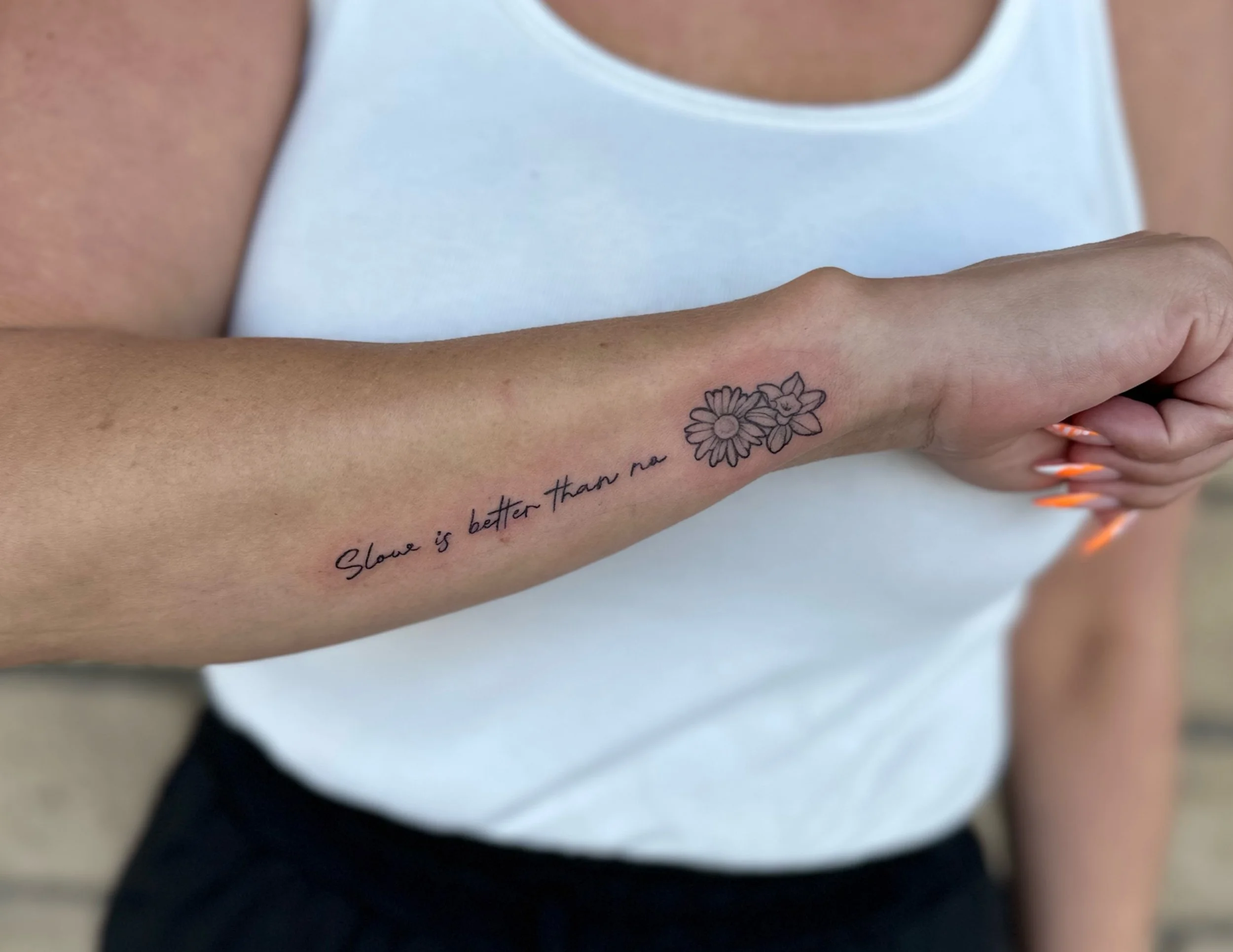 A person's arm with a tattoo of a flower and cursive text, wearing a white sleeveless shirt, with part of their hand visible.