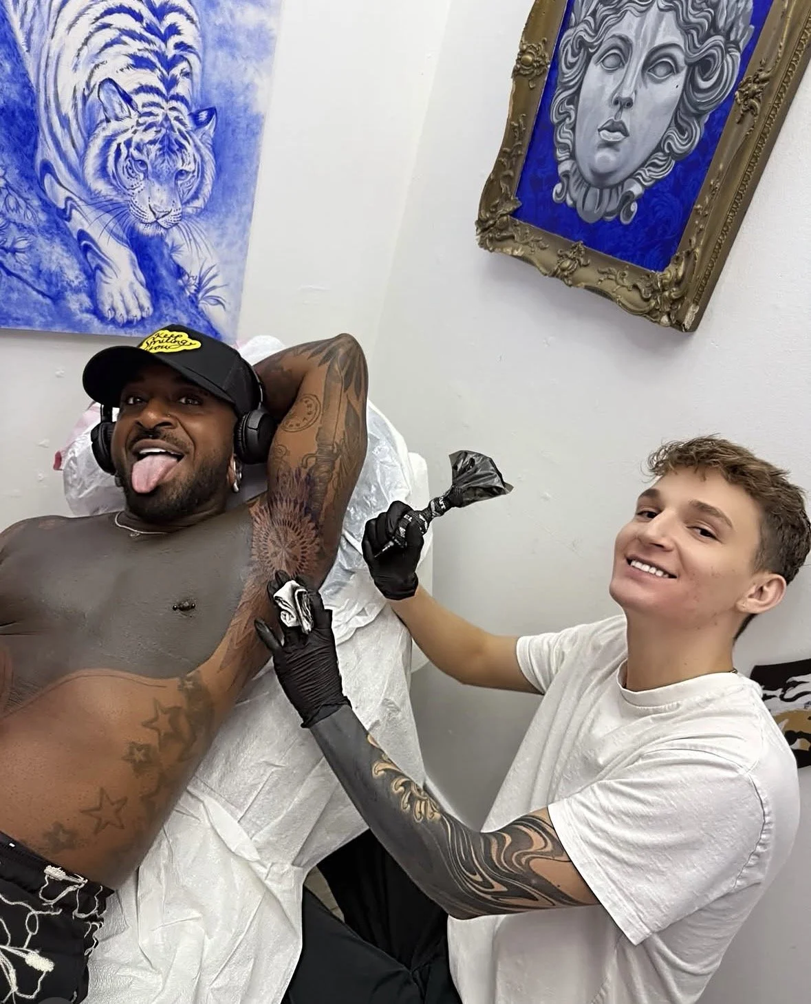 A man with tattoos lying on a medical chair, sticking out his tongue and smiling, wearing headphones and a cap. Another young man with tattoos on his arm is tattooing him, wearing black gloves and holding a tattoo machine. The scene is indoors, with blue artwork of a tiger and a classical face on the white wall behind them.
