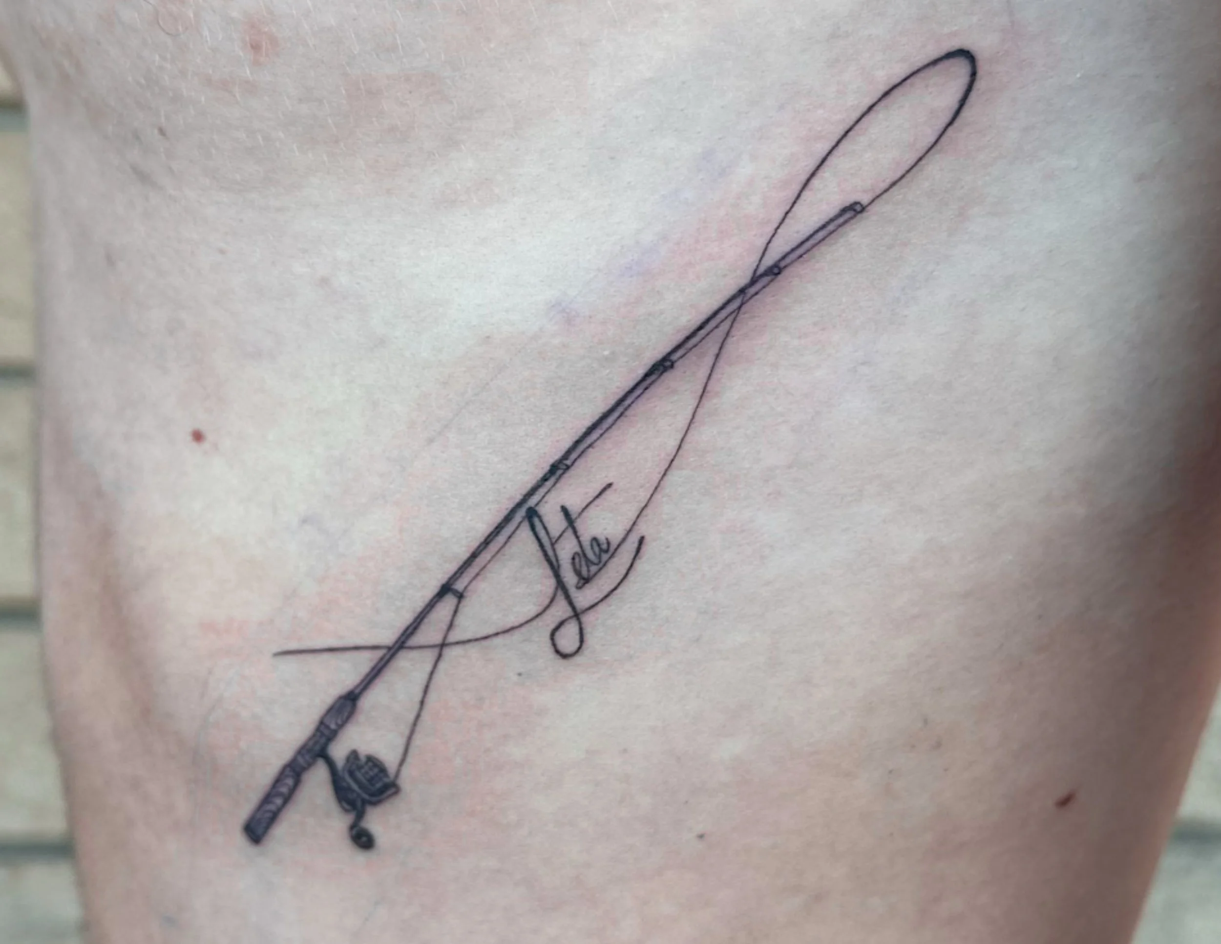 Close-up of a tattoo of a needle with a cord, with the word 'Vola' written below, on skin.