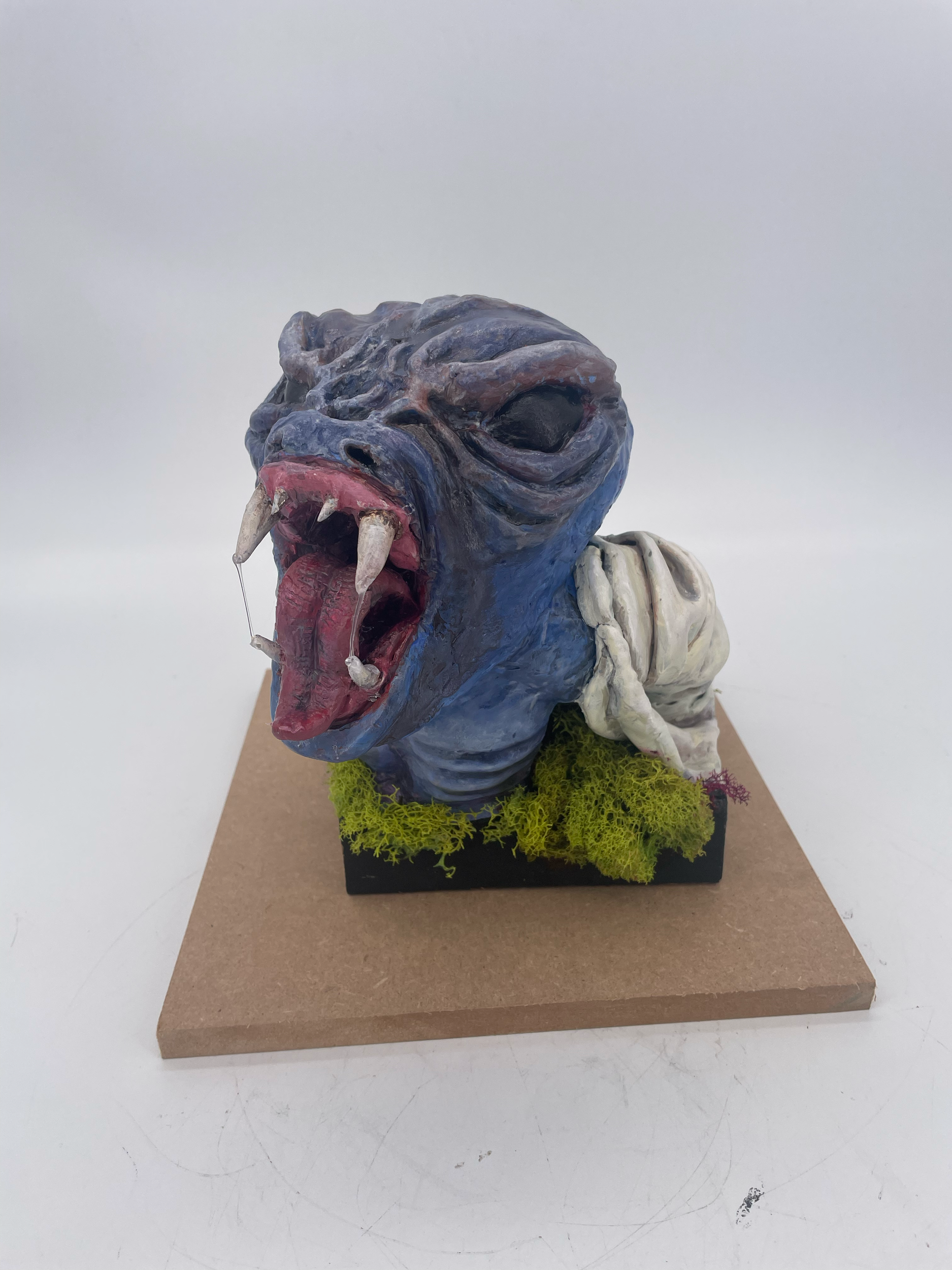 A sculpture of a mythical creature with a blue face, open mouth revealing sharp teeth and a pink tongue, and closed eyes, set on a black base with greenery and moss.