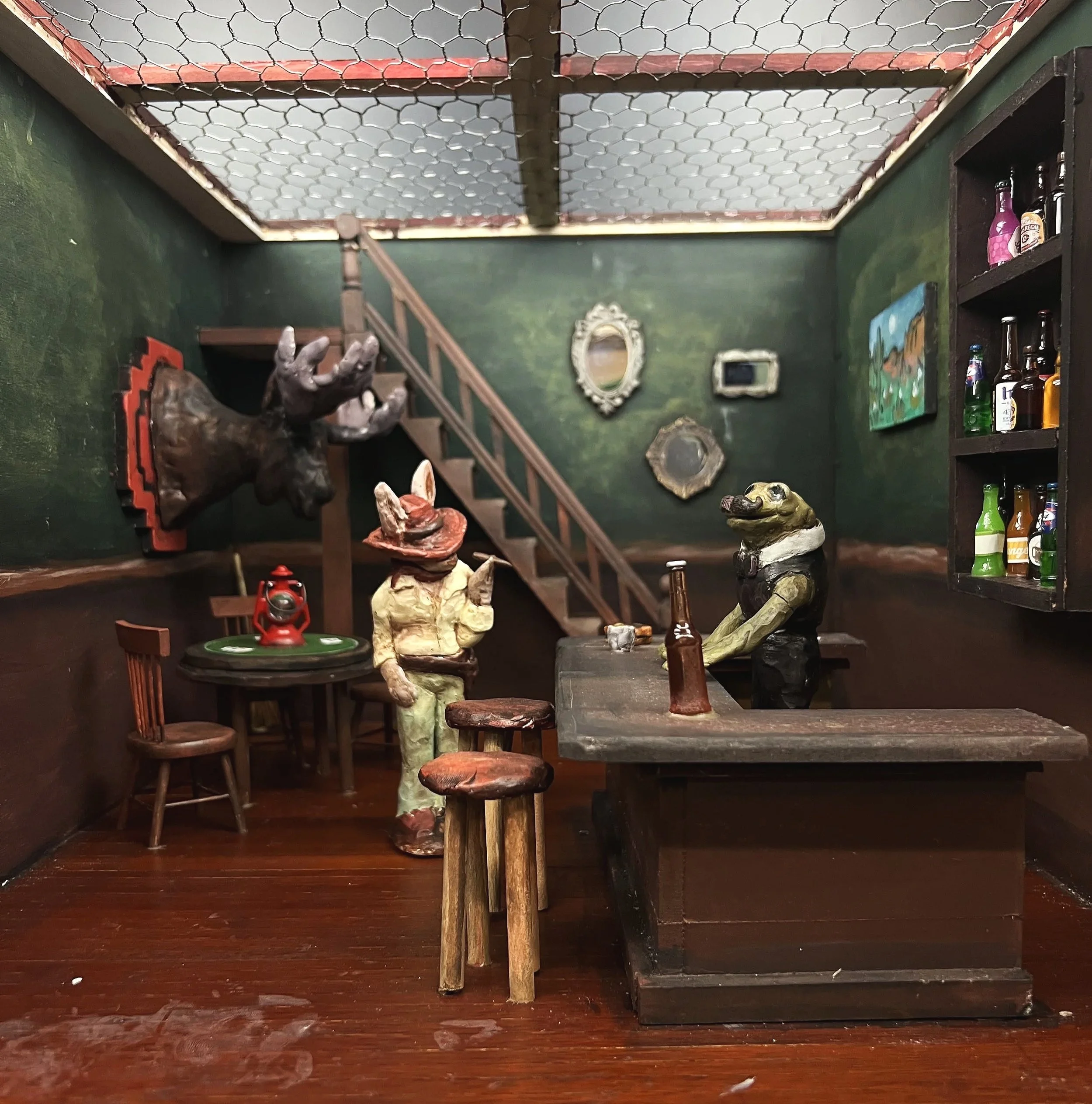 Miniature scene of a bar with animal characters, including a crocodile bartender and two human-like figures, in a cozy, decorated room with mirrors and bottles on shelves.