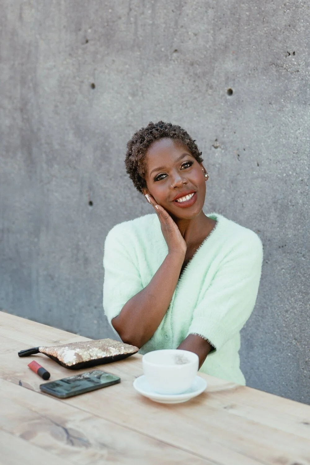 A woman with short curly hair and dark skin, smiling and touching her face, sitting at a wooden table with a cup of coffee, a sequin pouch, a lipstick, and a smartphone, in front of a gray concrete wall.