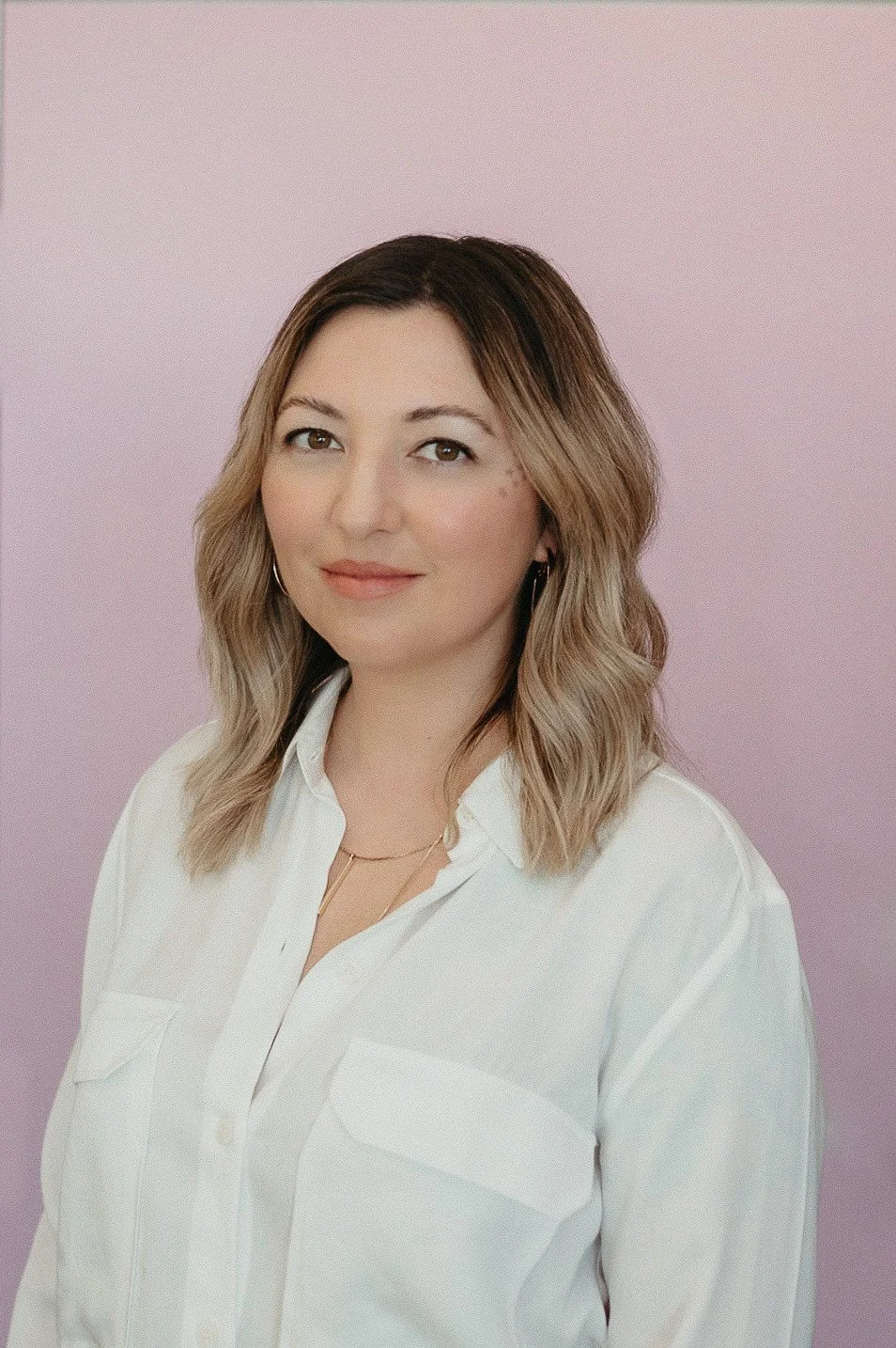 A woman with shoulder-length wavy hair wearing a white button-up shirt and layered necklaces, standing against a soft pink background.