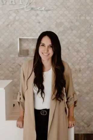 A woman with long dark hair smiling, wearing a beige coat over a white top and black pants, in front of a textured wall.
