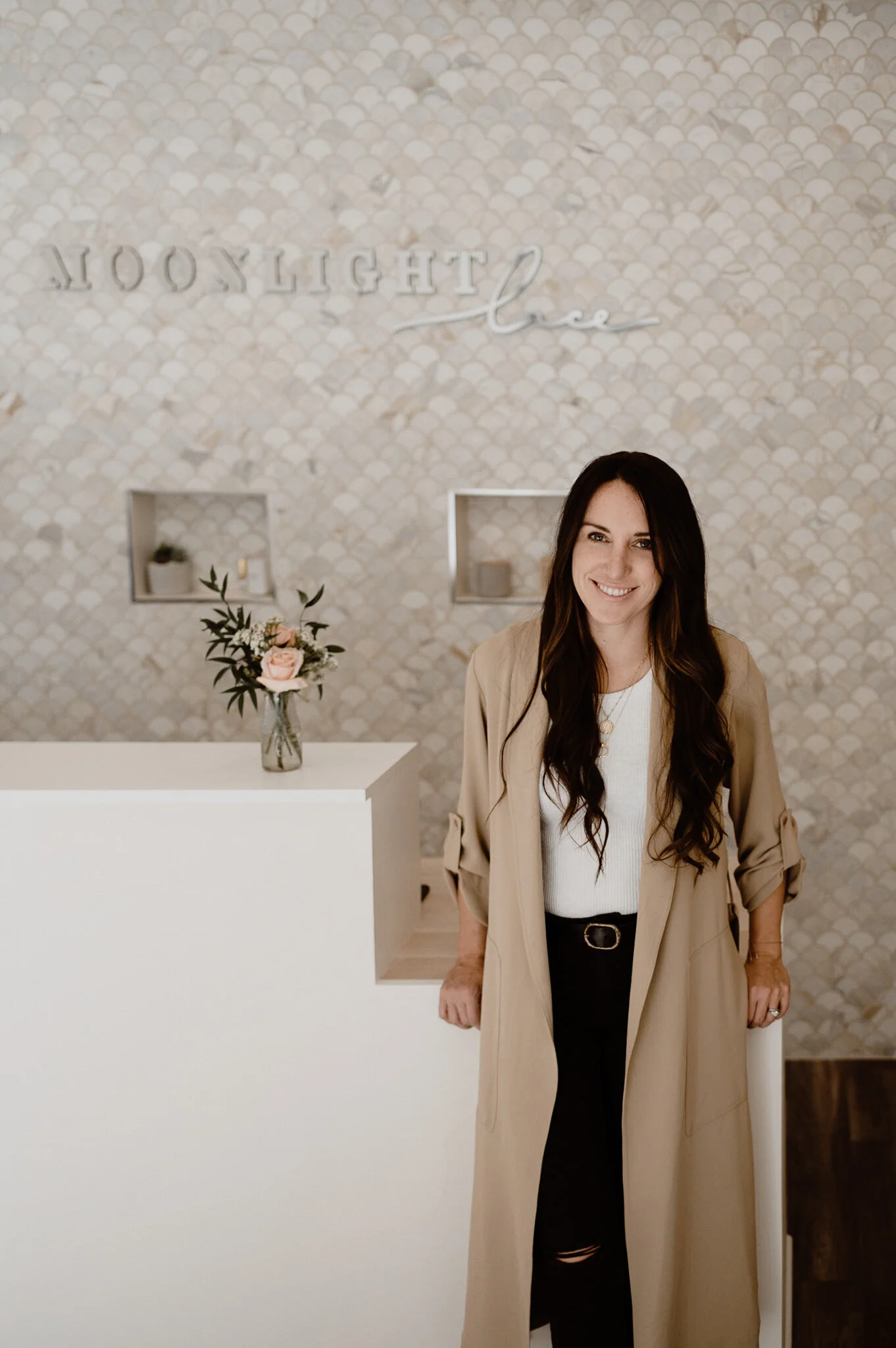 A woman with long dark brown hair, wearing a light beige coat over a white top and black pants, standing inside a modern home with a patterned wall that reads 'LITTLE,