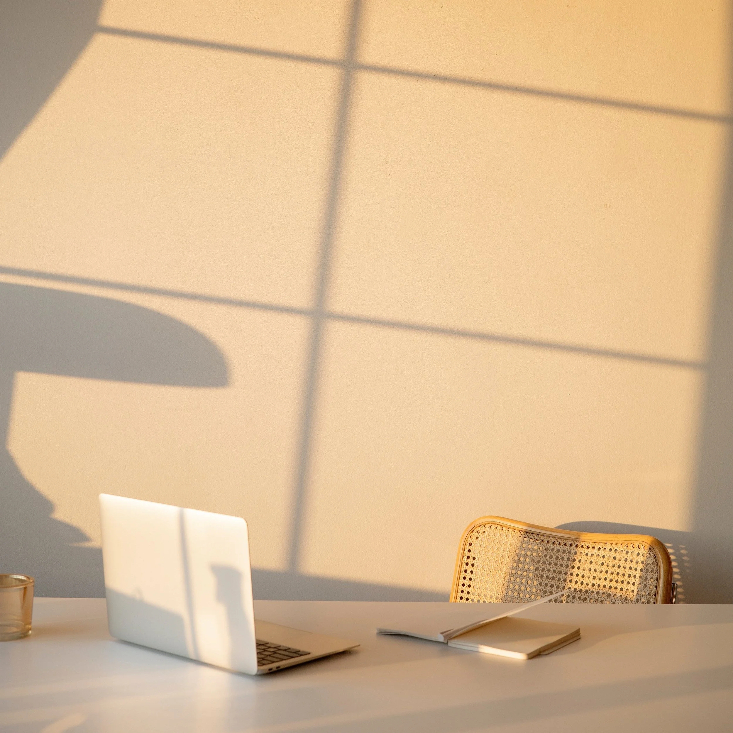 A desk with a closed laptop, a white plate, a notebook, a glass, and a wicker chair, illuminated by sunlight creating shadows on the beige wall.