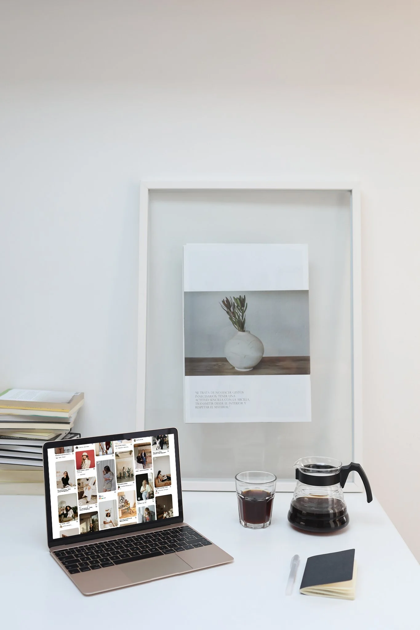 A laptop displaying a social media photo grid on a white desk, with a glass of iced coffee, a glass of black coffee, a black notebook with a pen, a small notebook, a stack of books, and a coffee pot. In the background, there is a white wall with a framed art piece featuring a white vase with green branches.