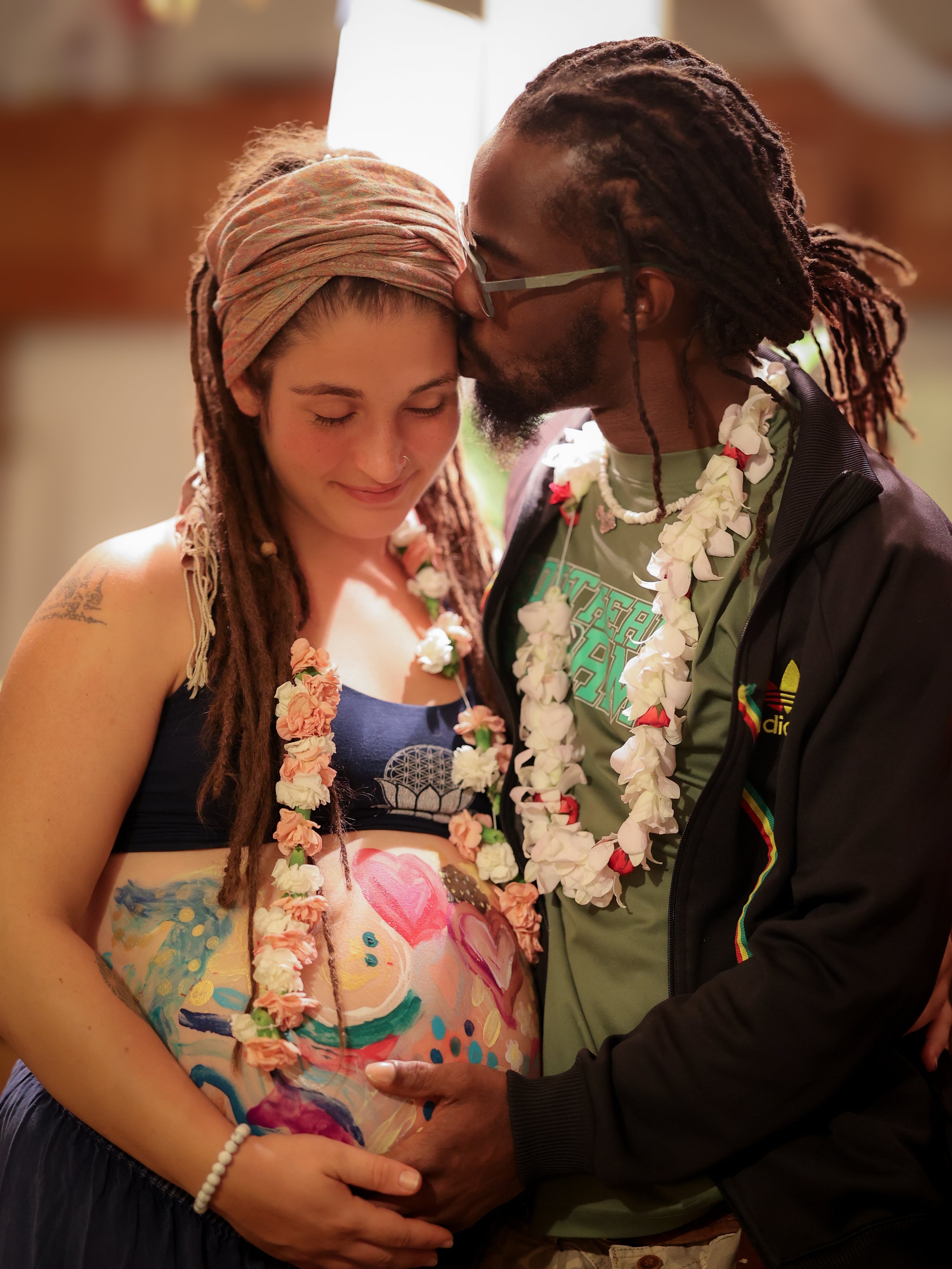 A pregnant woman with painted belly featuring colorful hearts and swirls, wearing a flower lei and with a headscarf, and a man with dreadlocks and glasses, wearing a necklace and a jacket, gently holding her pregnant belly. They are in an intimate mo