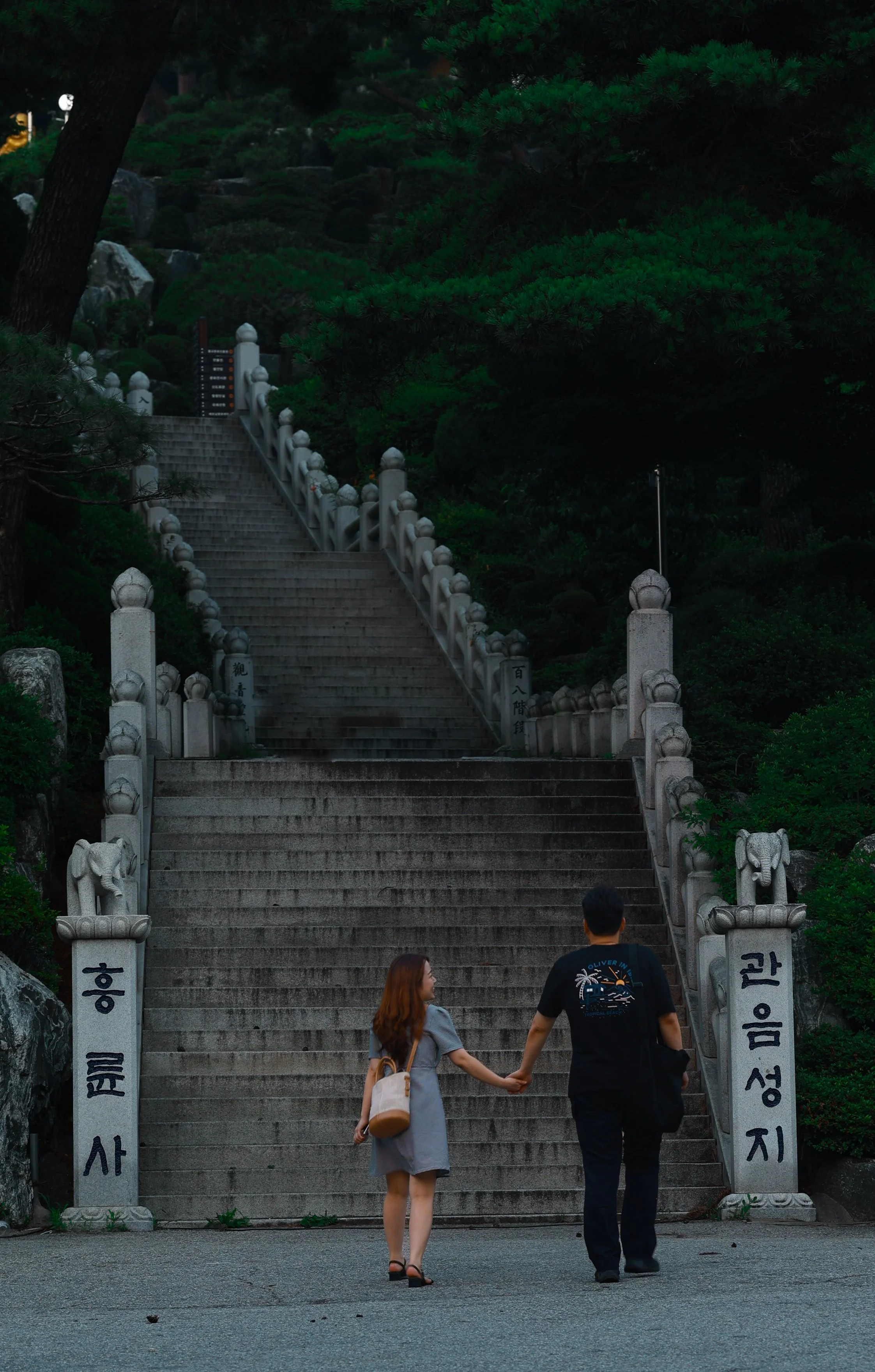 A man and woman holding hands at the base of a long stone staircase surrounded by lush green trees, with traditional Korean stone pillars at the entrance.