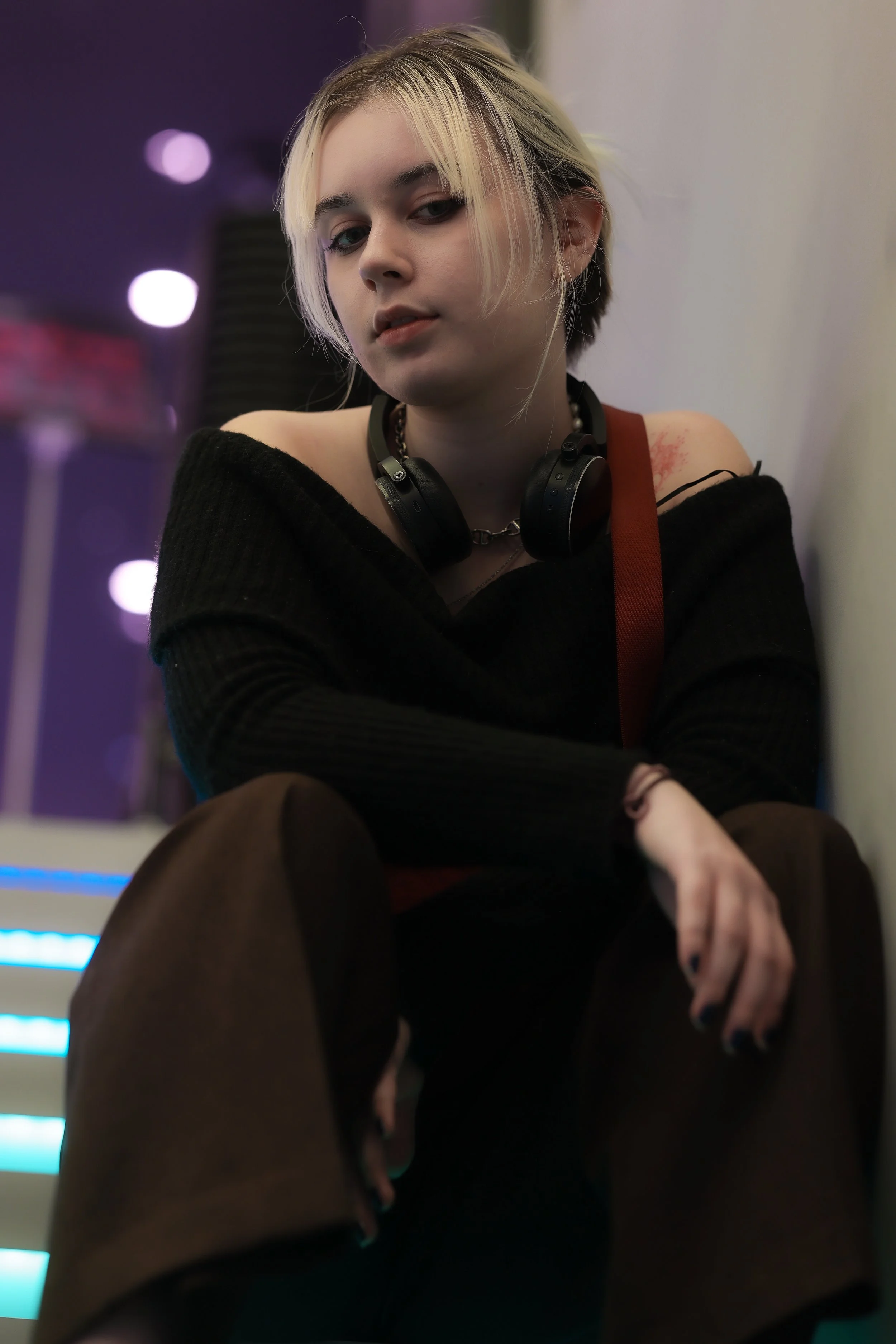 A young woman with short, bleached blonde hair and dark eye makeup, wearing a black off-shoulder sweater, headphones around her neck, sitting with a relaxed posture in an indoor setting with purple and white lighting.
