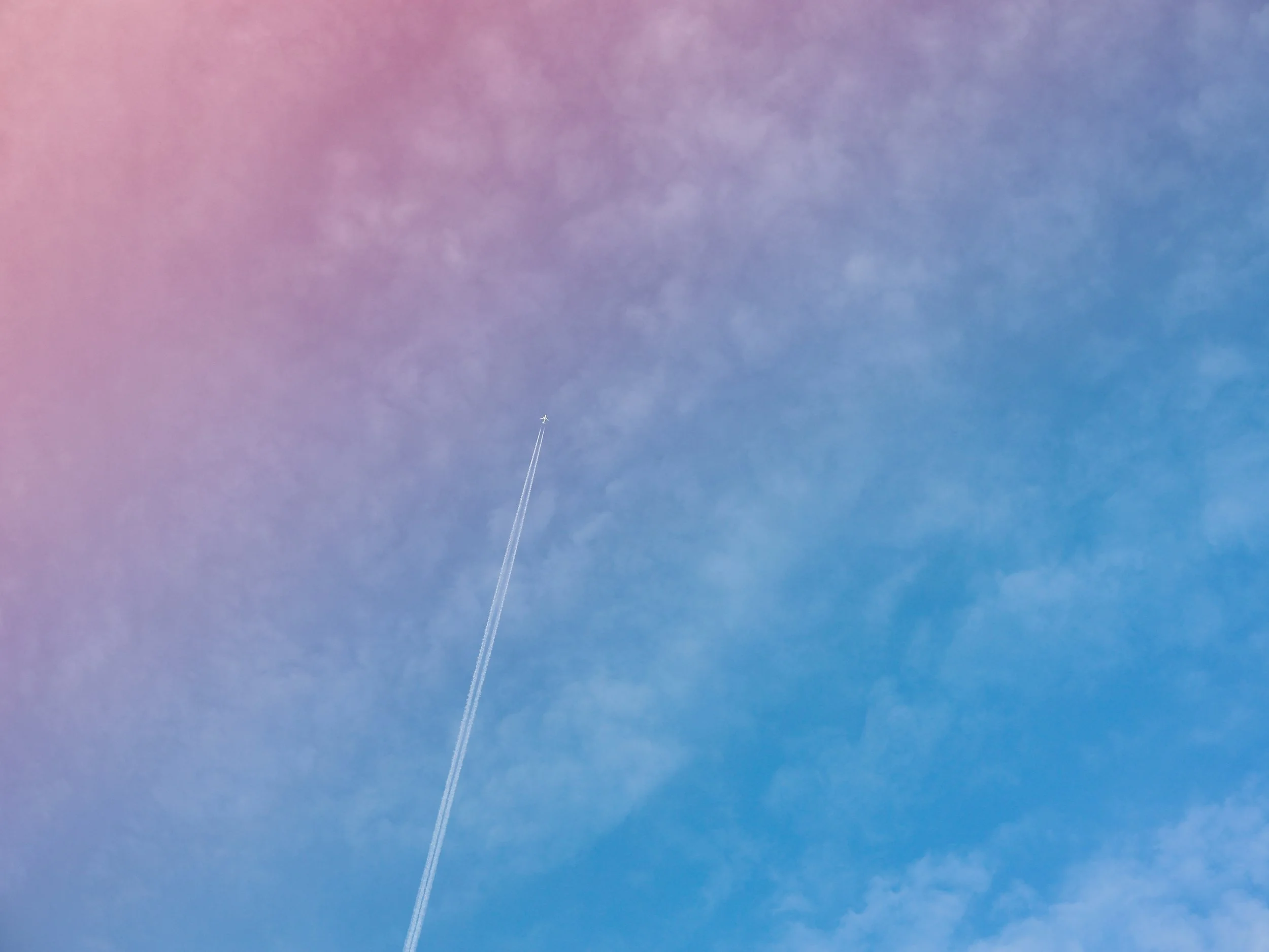The sky with clouds that have a pinkish-purple hue and a small airplane in the distance, leaving a contrail behind.