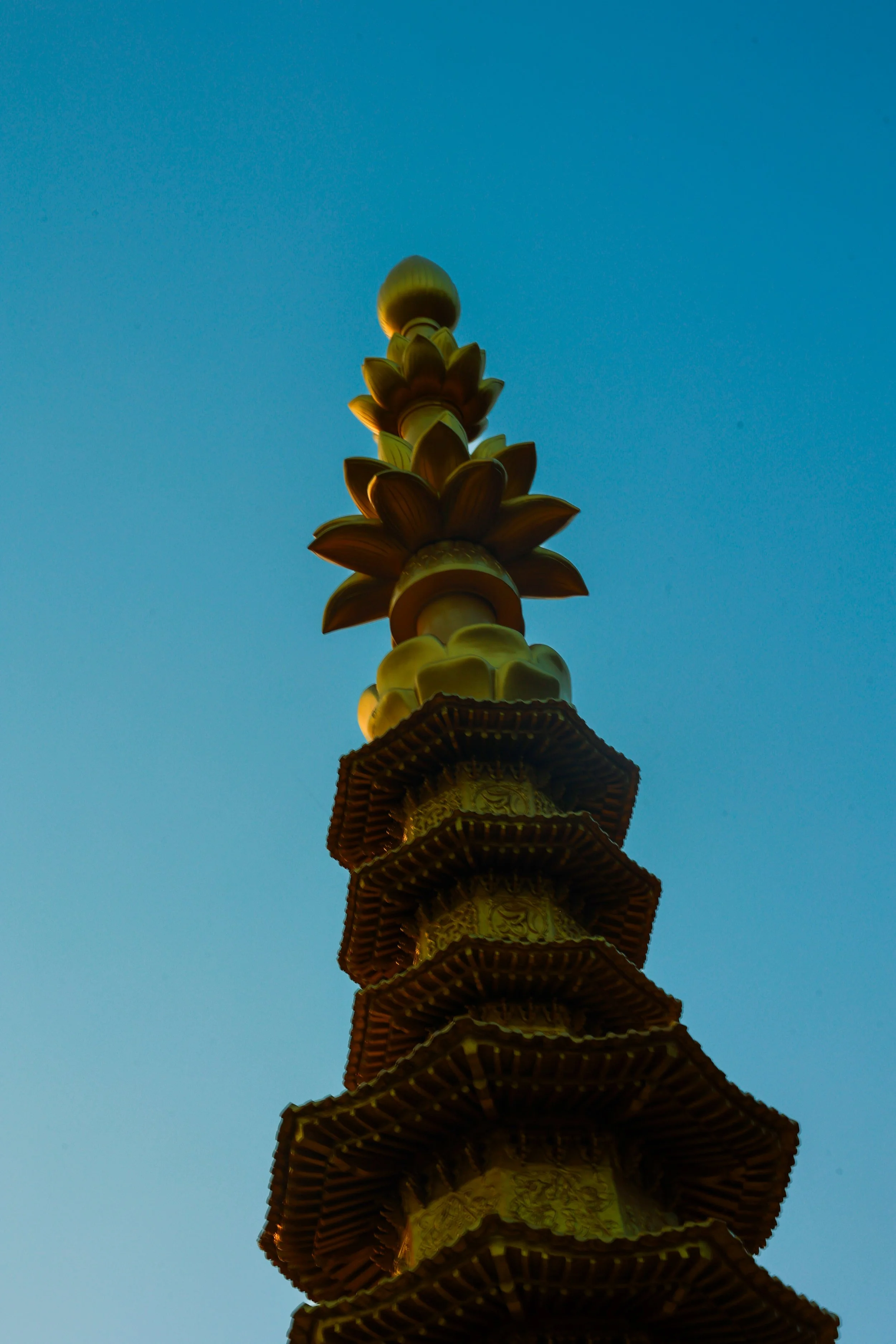 View upward of a tall, ornate tower with decorative floral and geometric patterns, topped with a rounded finial, set against a clear blue sky.