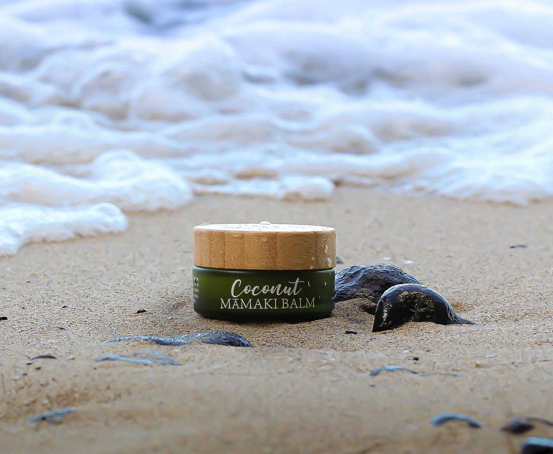A container of coconut MAMAKI BALM sits on sandy beach near rocks with ocean waves in the background.