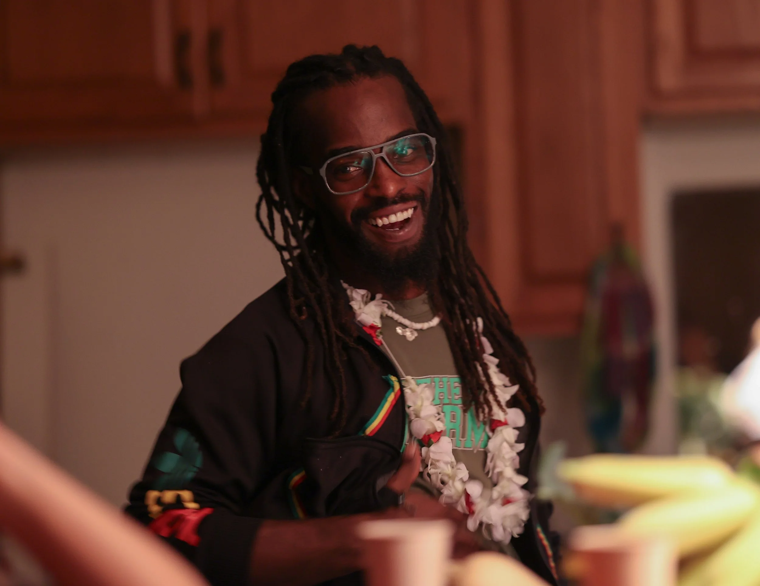 A man with dreadlocks wearing glasses and a lei, smiling at an indoor gathering.