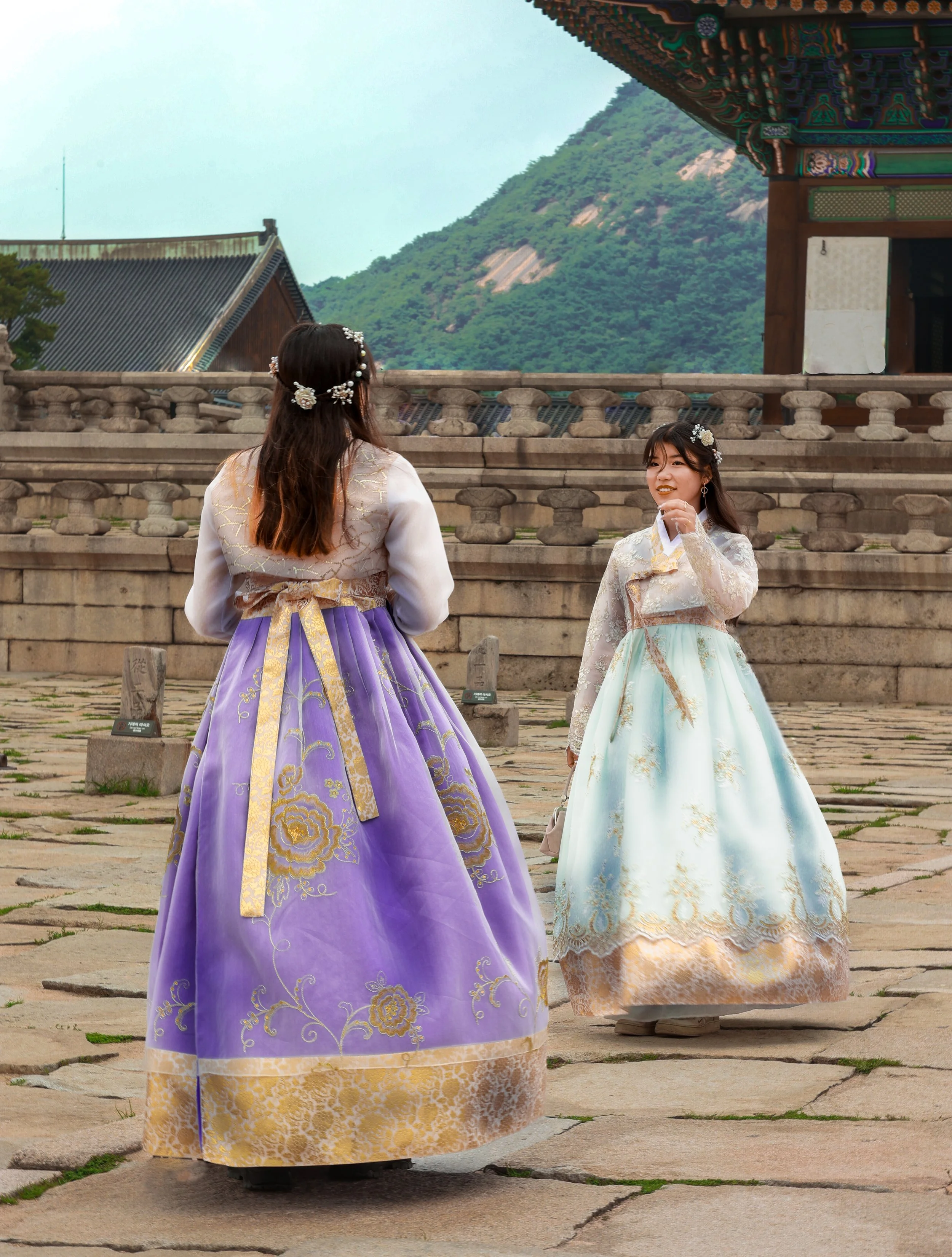 Two women wearing traditional Korean hanbok dresses with floral hair accessories standing outdoors in front of stone steps and a colorful building, with green mountains in the background.